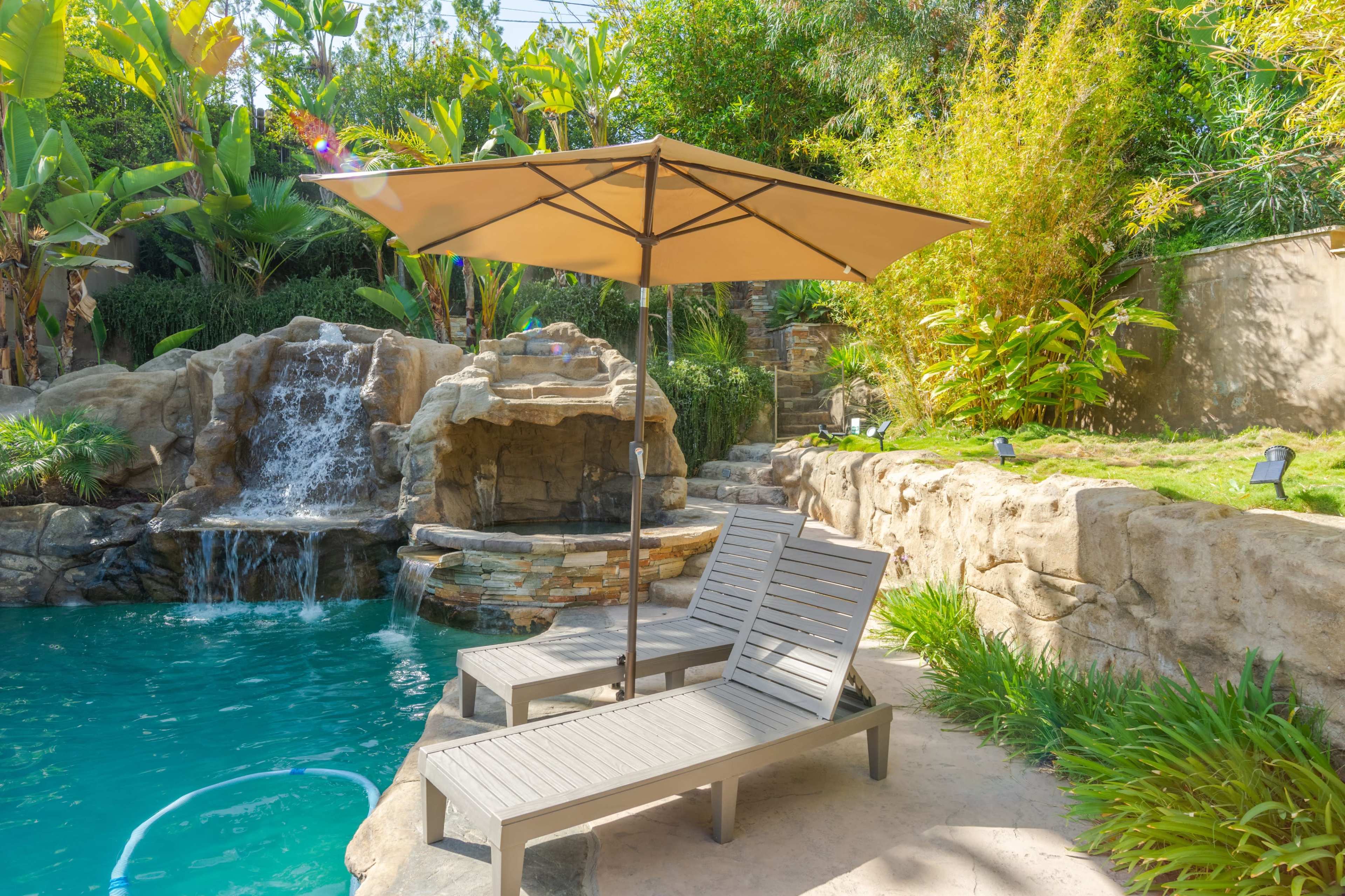 A tranquil poolside setting with two wooden lounge chairs positioned beneath a beige umbrella, adjacent to a waterfall and surrounded by lush greenery.
