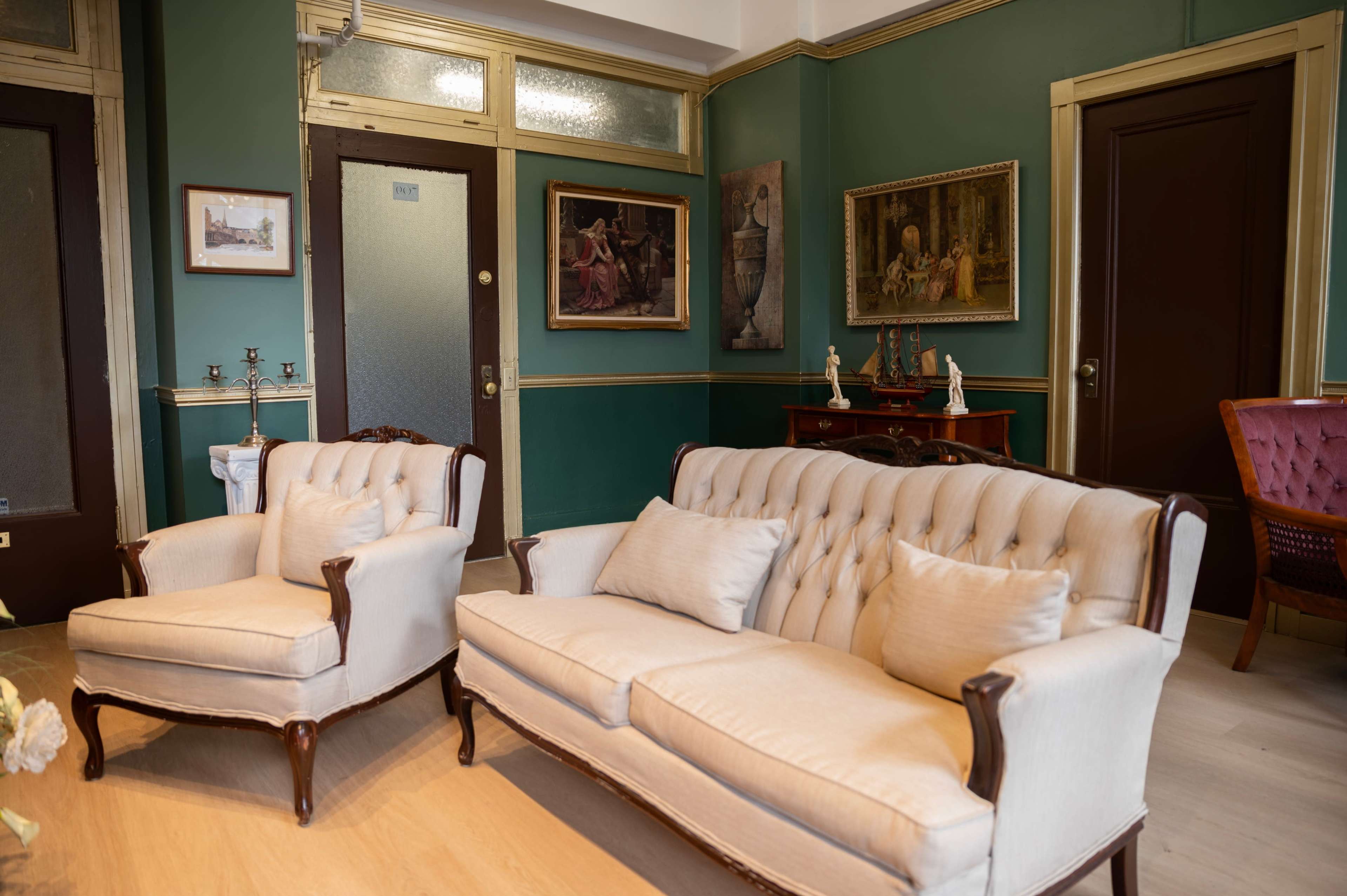 A vintage living room with two upholstered armchairs and a tufted sofa, framed paintings on the walls, and a decorative wooden cabinet.