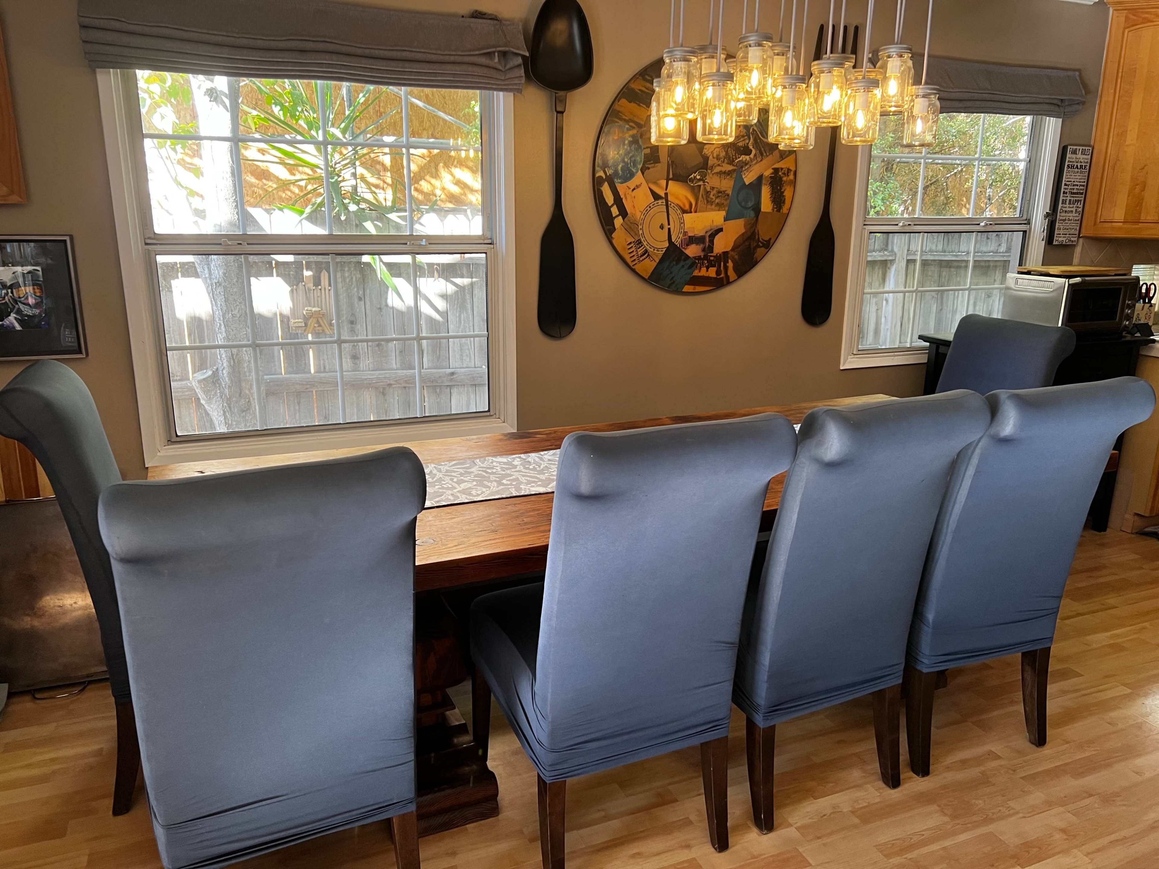 A wooden dining table with high-backed blue chairs is set in a well-lit room featuring large windows and a decorative circular wall piece above the table.