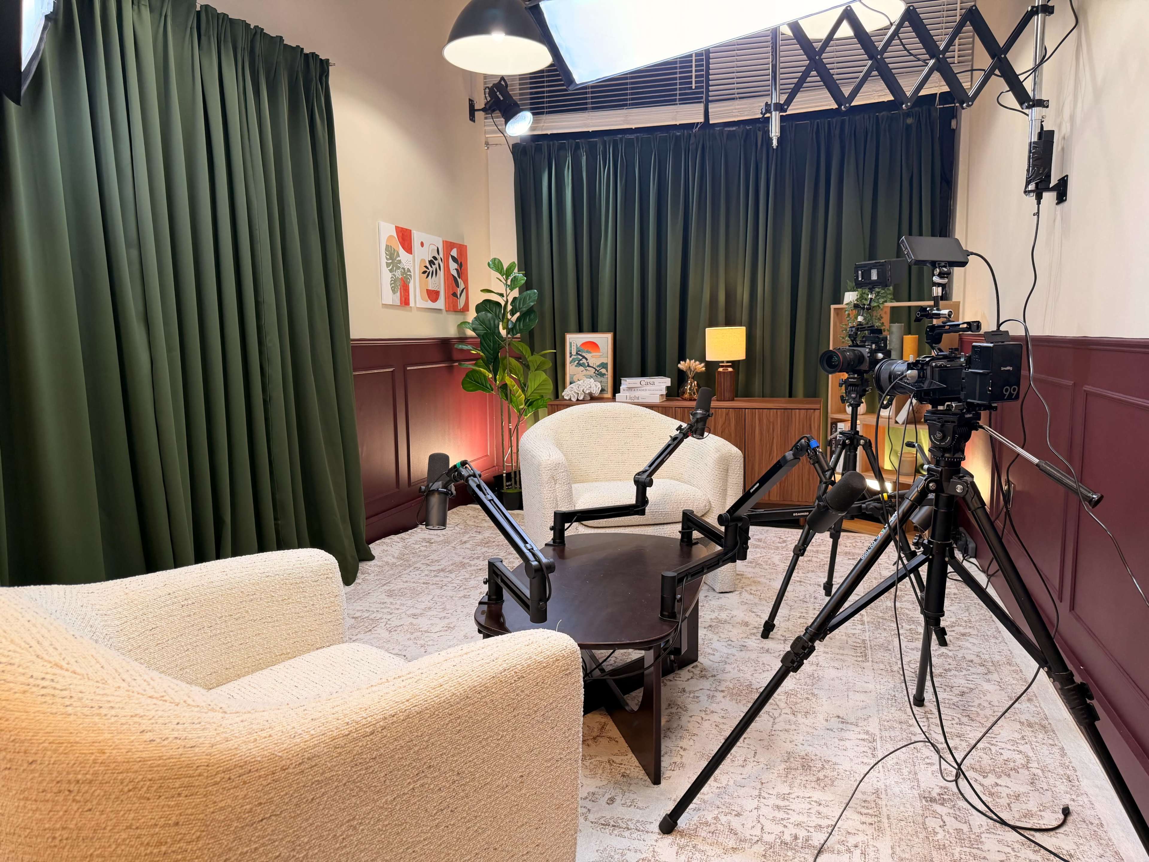 The image shows a cozy interview setup featuring two light-colored chairs facing a round table, surrounded by cameras and lighting equipment in a room with dark green curtains.