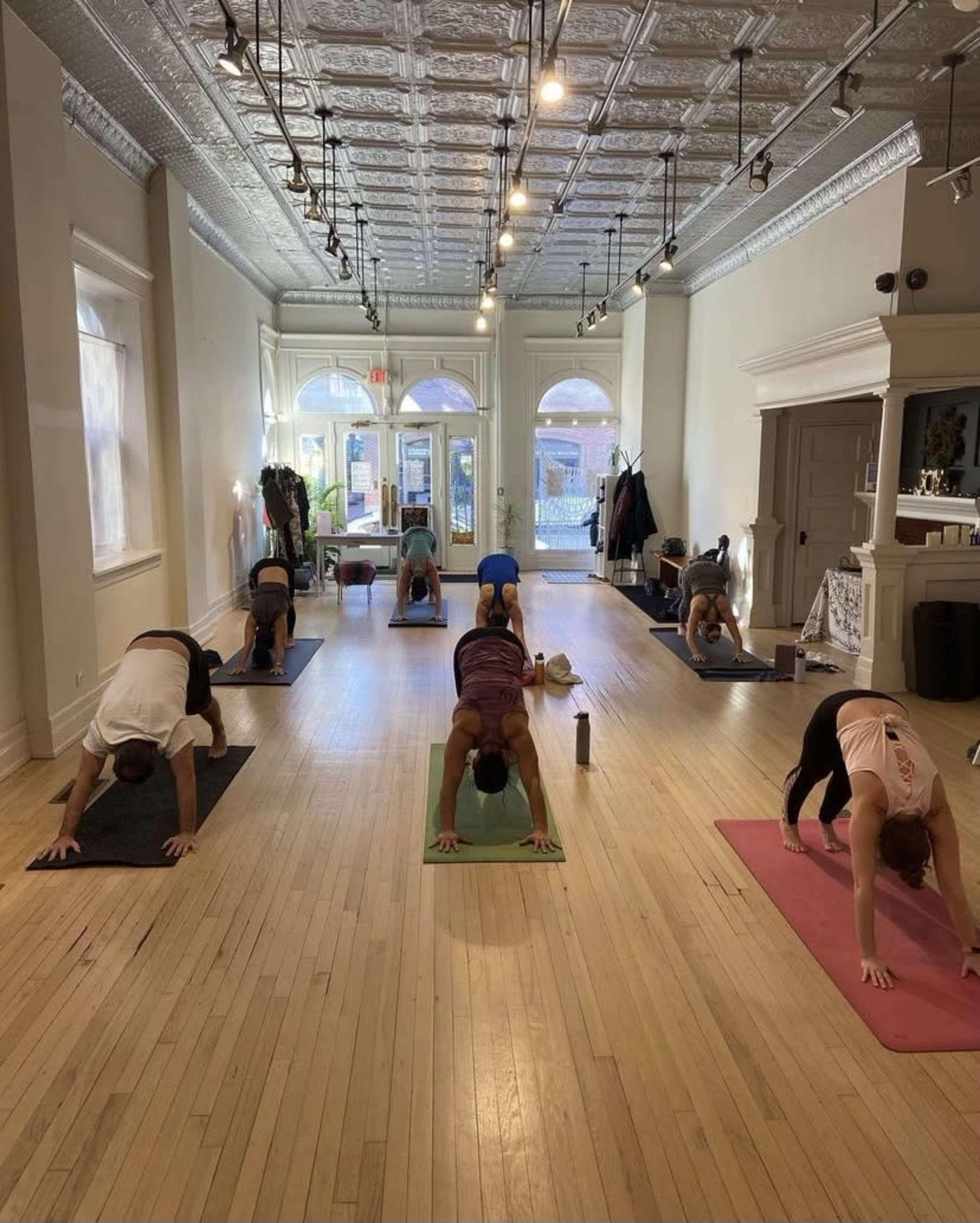 Several people participate in a yoga class, practicing downward dog poses on mats in a well-lit studio with wooden floors.