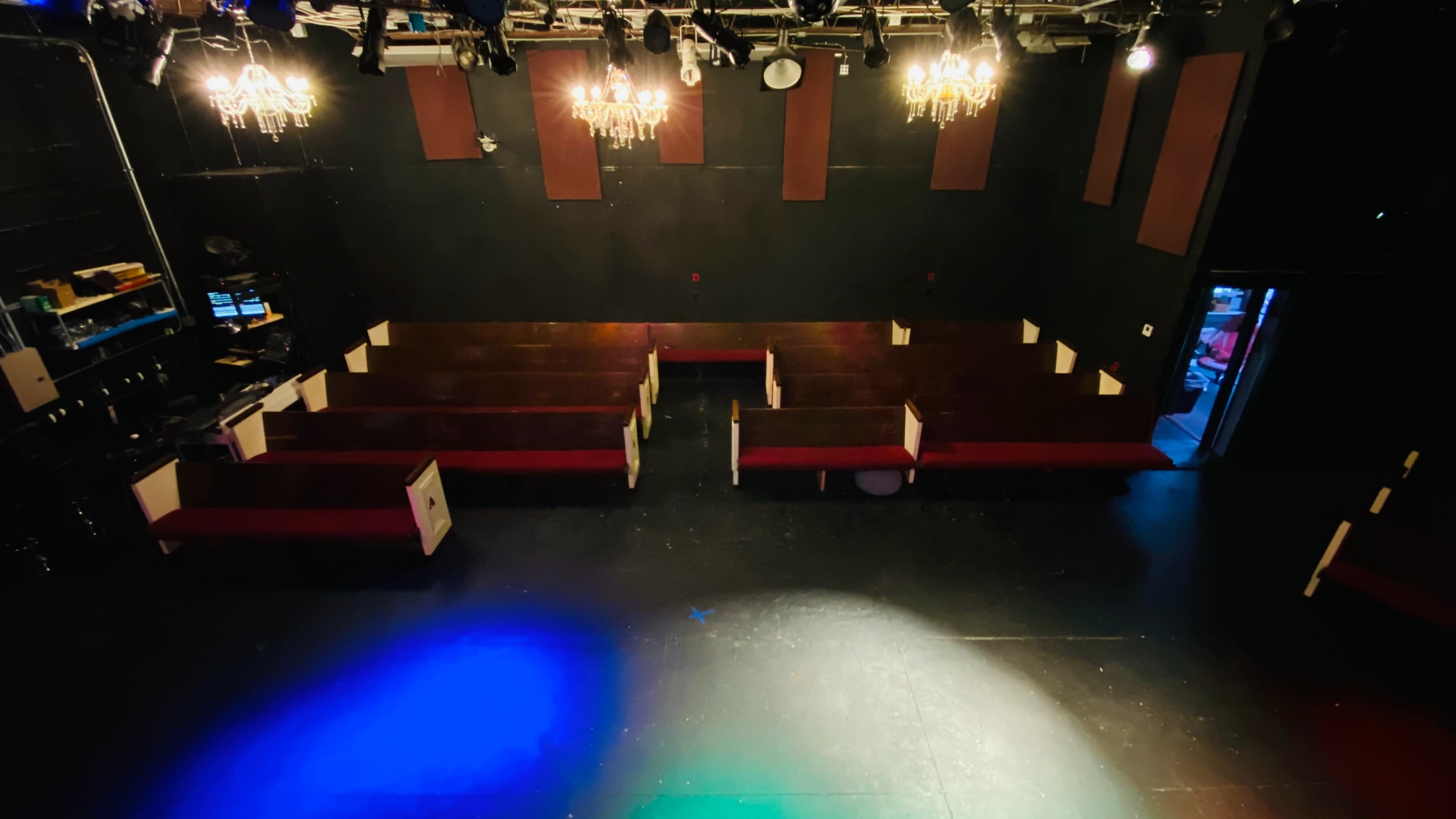 The image shows a theater stage with rows of wooden benches and chandeliers hanging from the ceiling, illuminated by colored lights.
