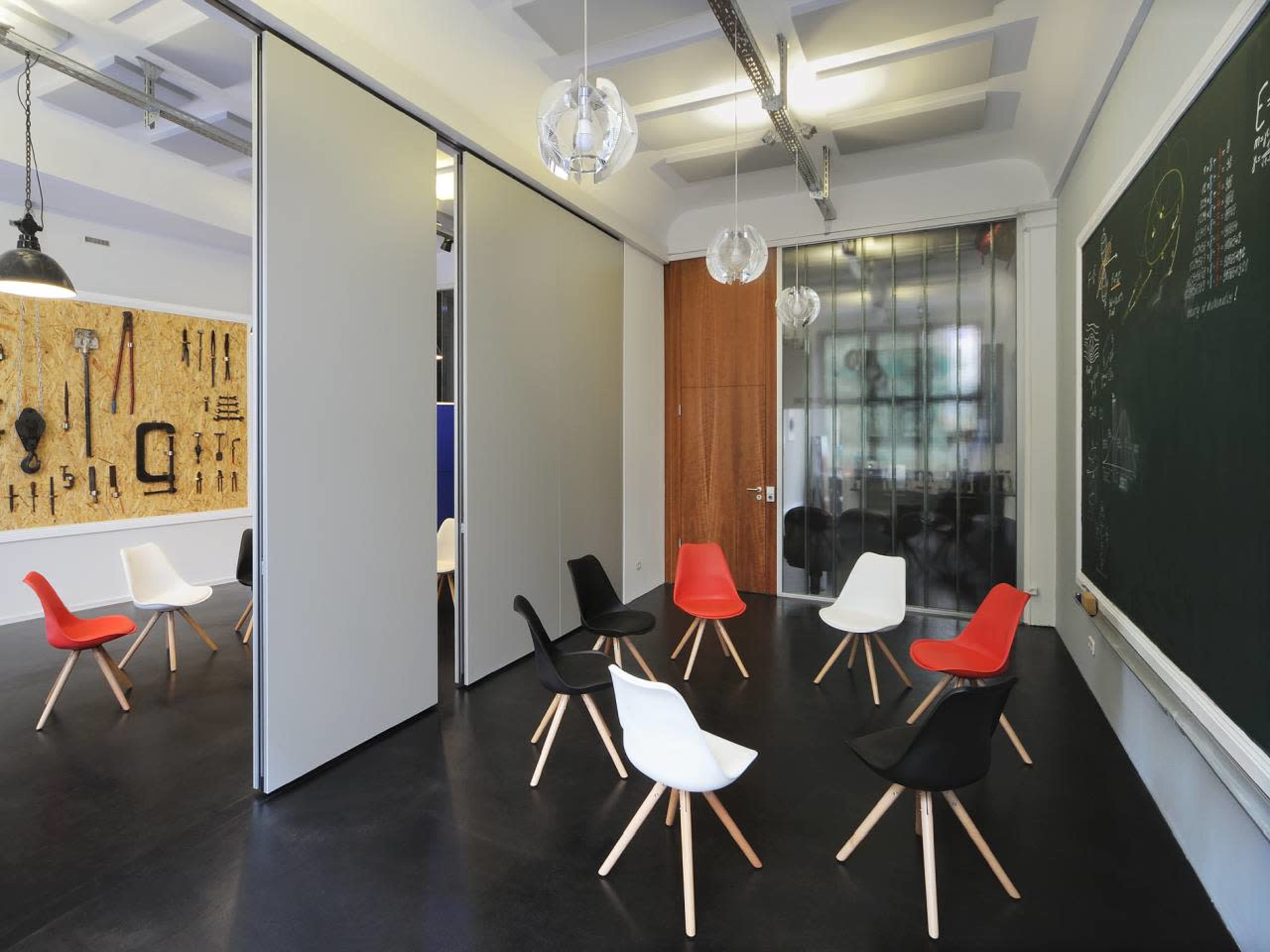 The image shows a minimalist meeting space with several chairs arranged on a black floor, surrounded by sliding partitions and a black chalkboard.