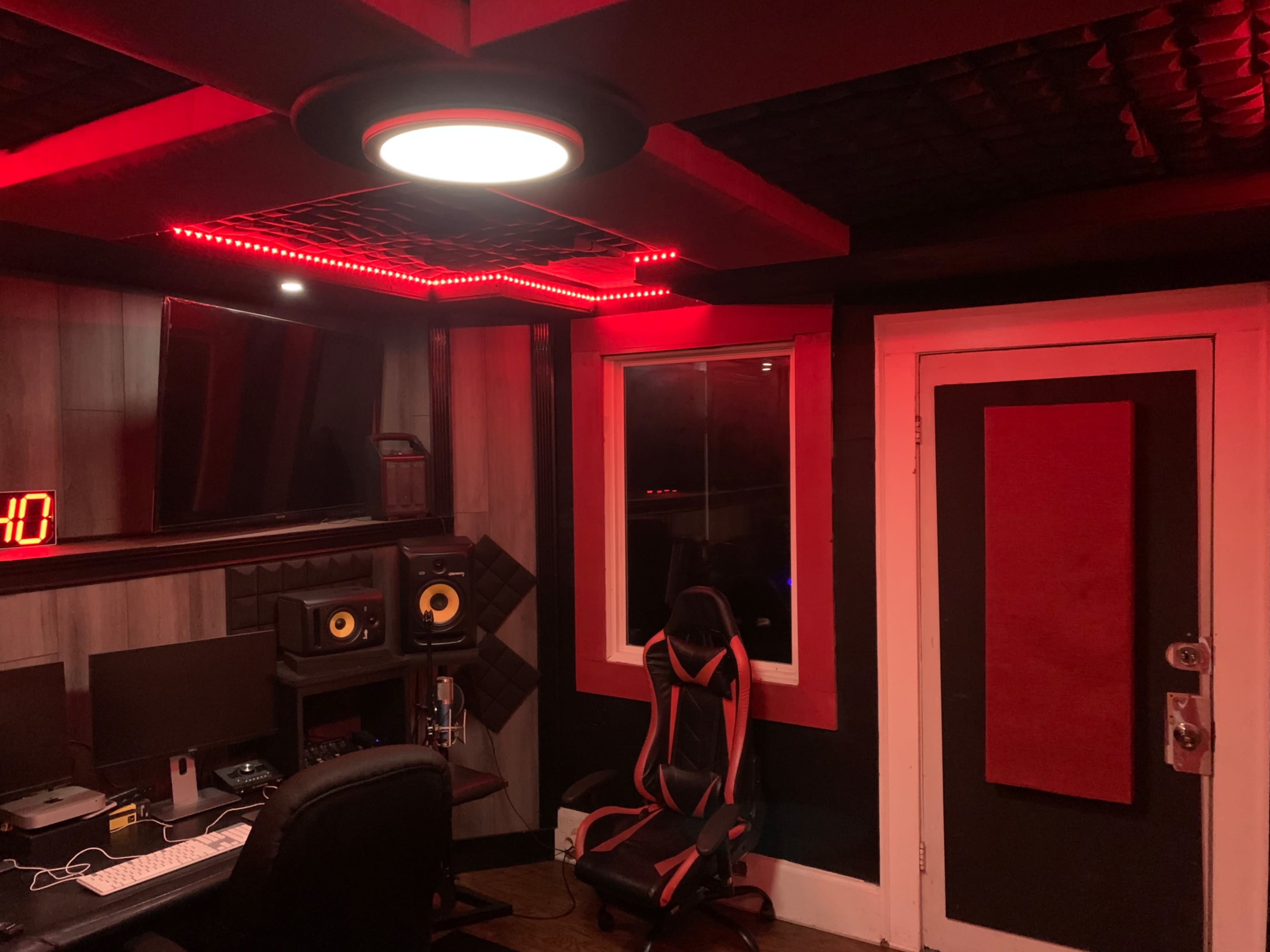 The image shows a music recording studio with red LED lighting, a computer desk, speakers, and a gaming chair.