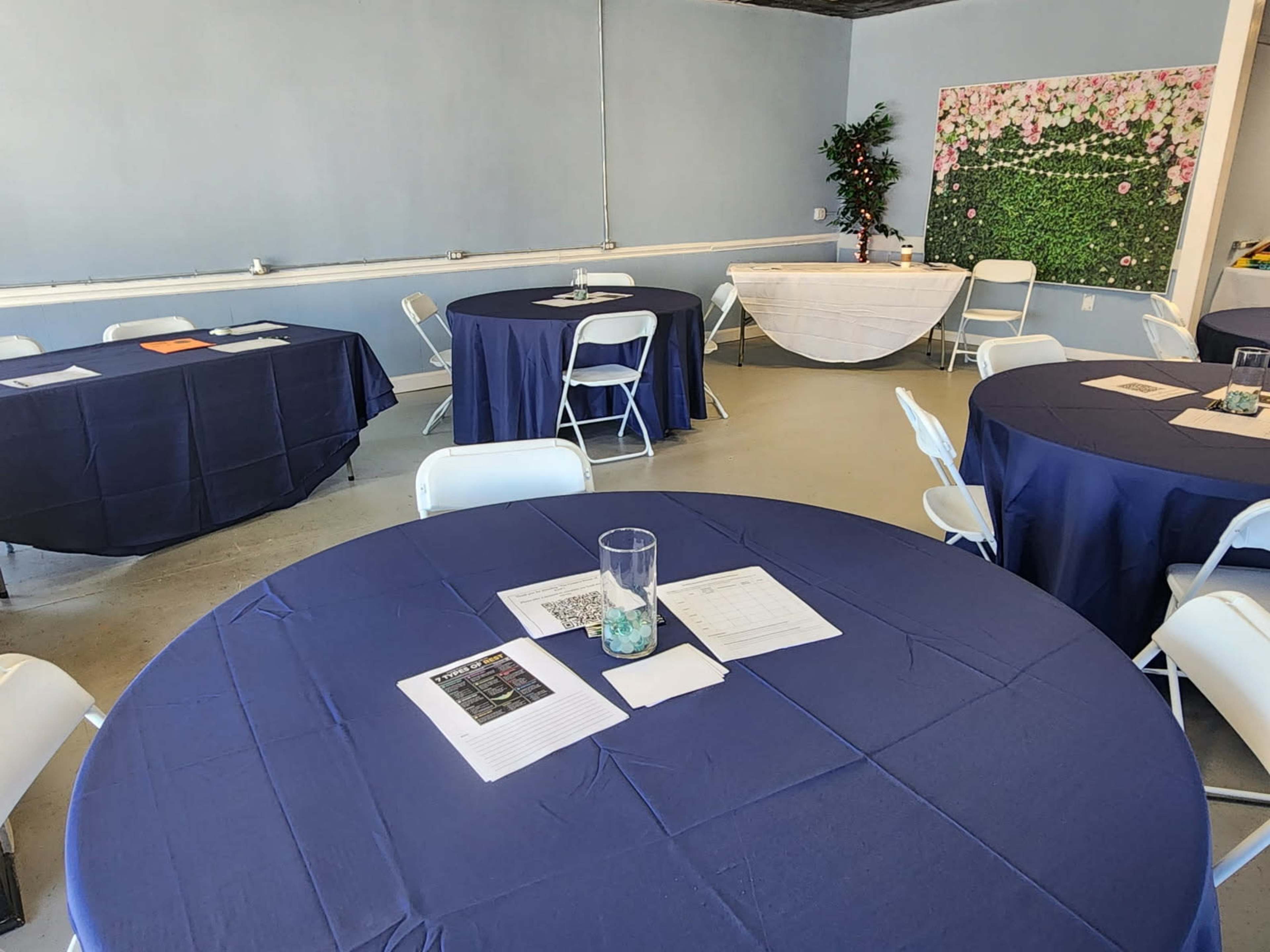 A spacious room features several round tables covered with navy blue tablecloths, each with documents and a glass vase in the center, alongside a decorated wall with flowers.