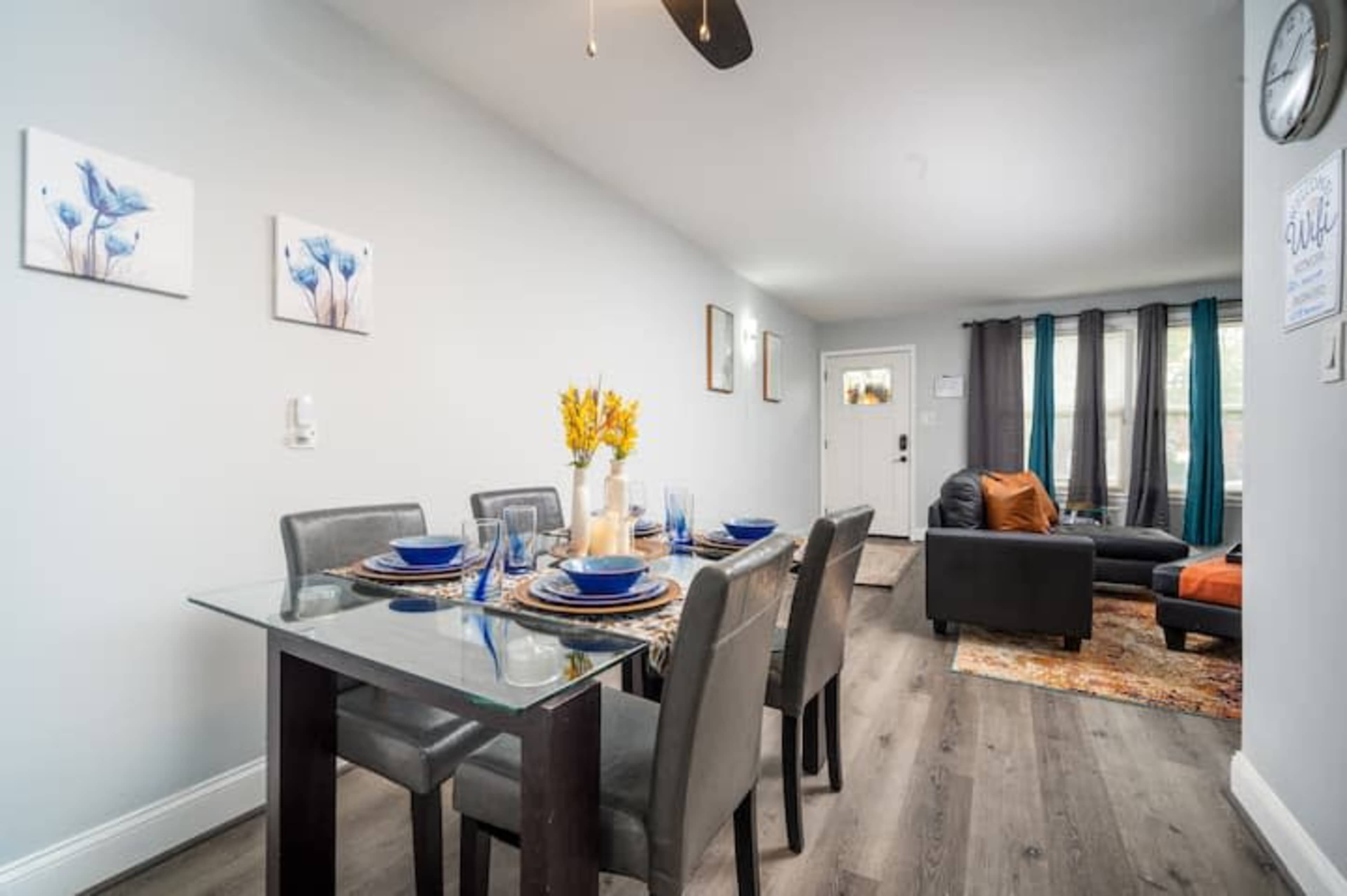 A dining area with a glass table set for six, surrounded by chairs, and connecting to a living room with a dark sofa and decorative elements.