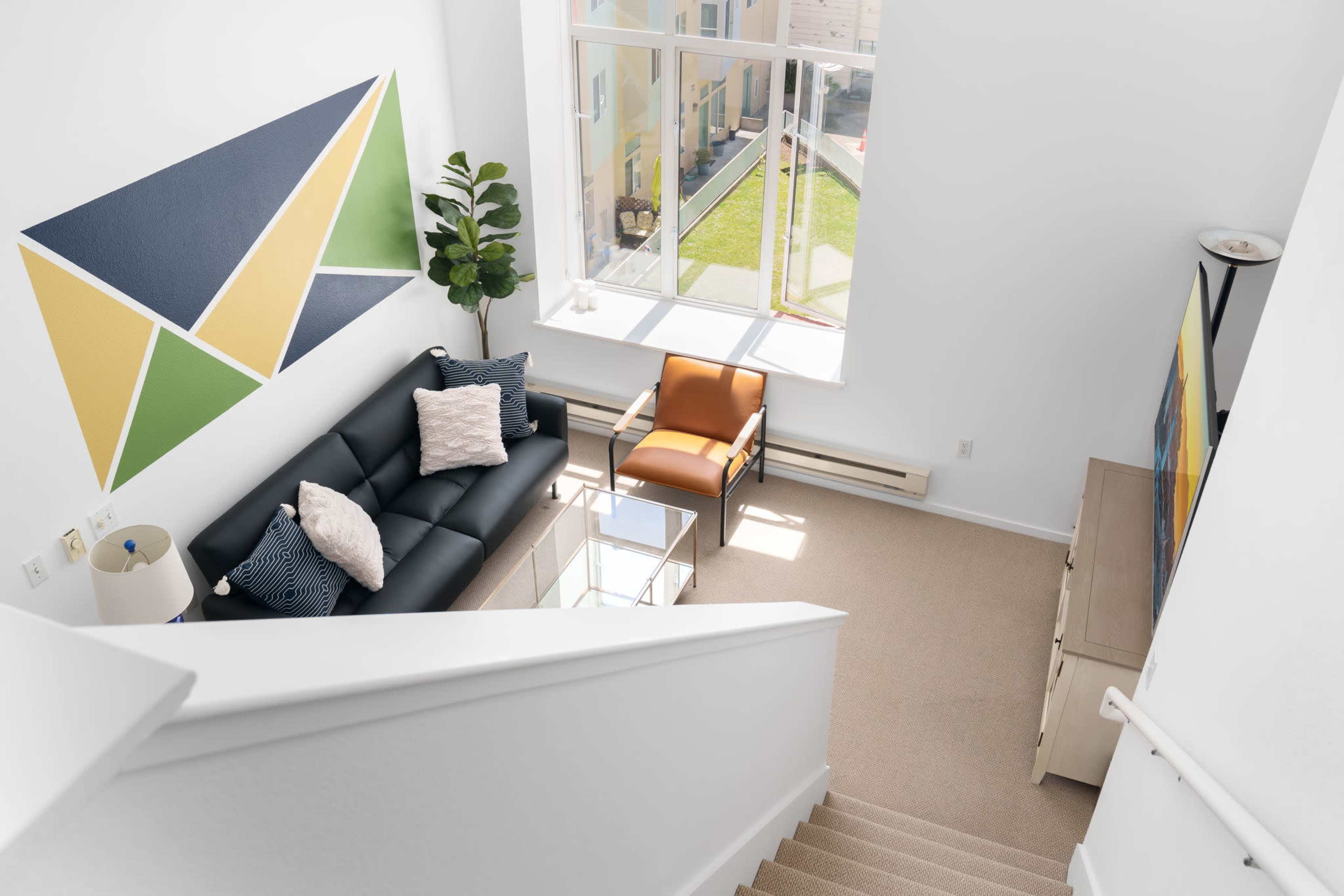 A modern living room with a black sofa, an orange armchair, a glass coffee table, and a large window offering a view of the outdoors.