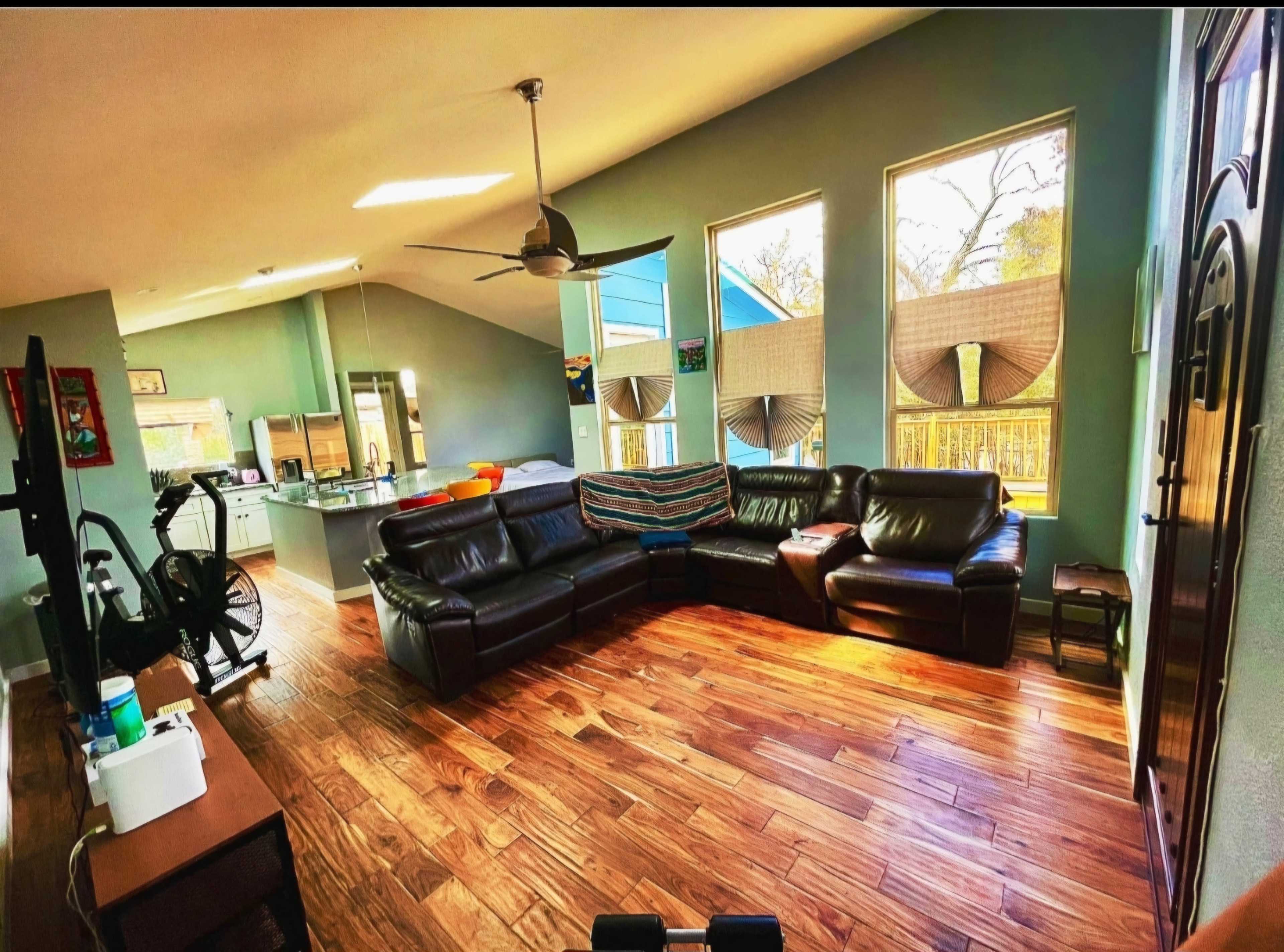The image shows a spacious living room with a leather sectional couch, large windows, wooden flooring, and a visible kitchen area in the background.