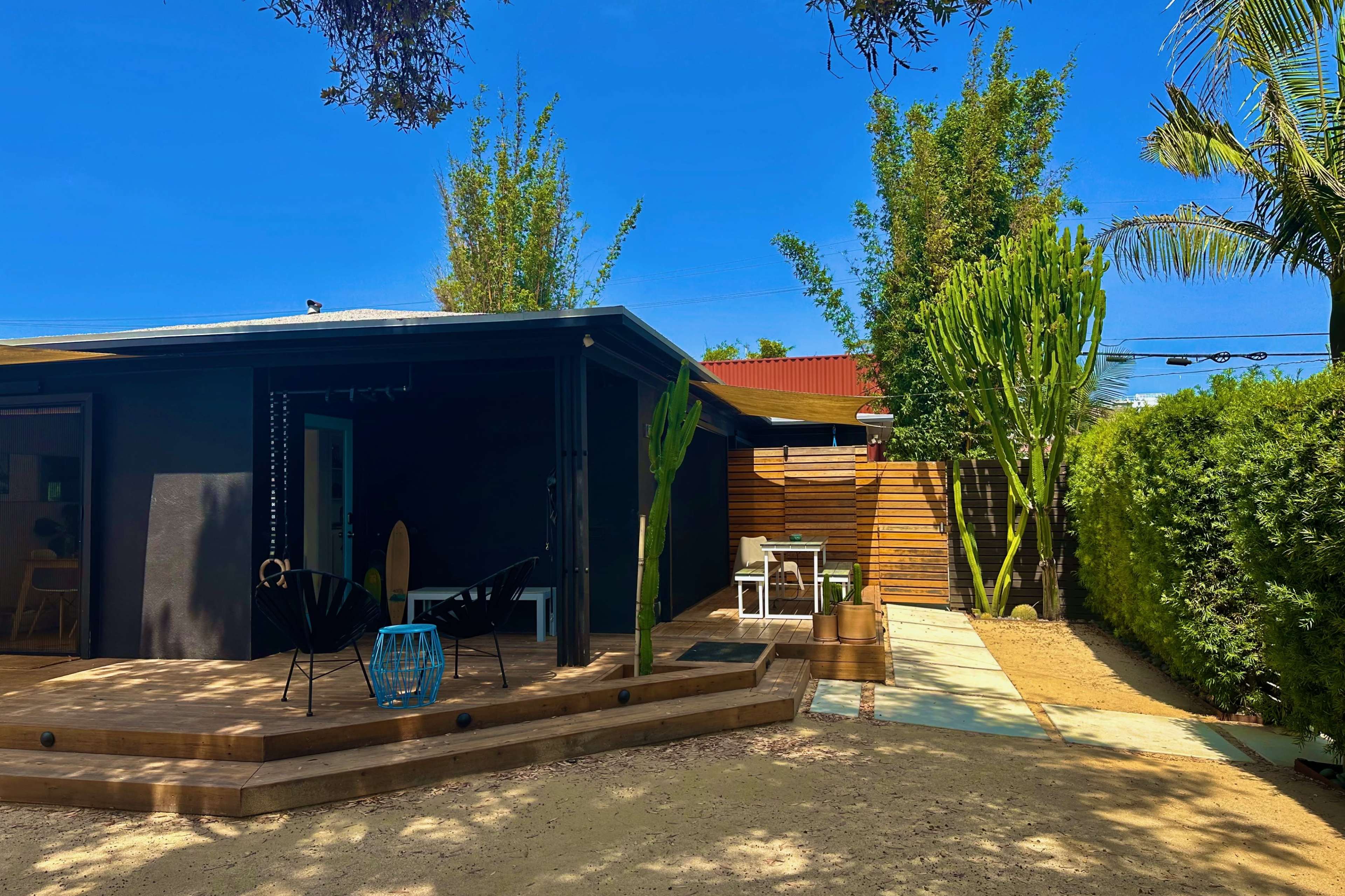 The image shows a modern outdoor space featuring a wooden deck with black chairs, cacti, and a pathway leading to a house surrounded by greenery.