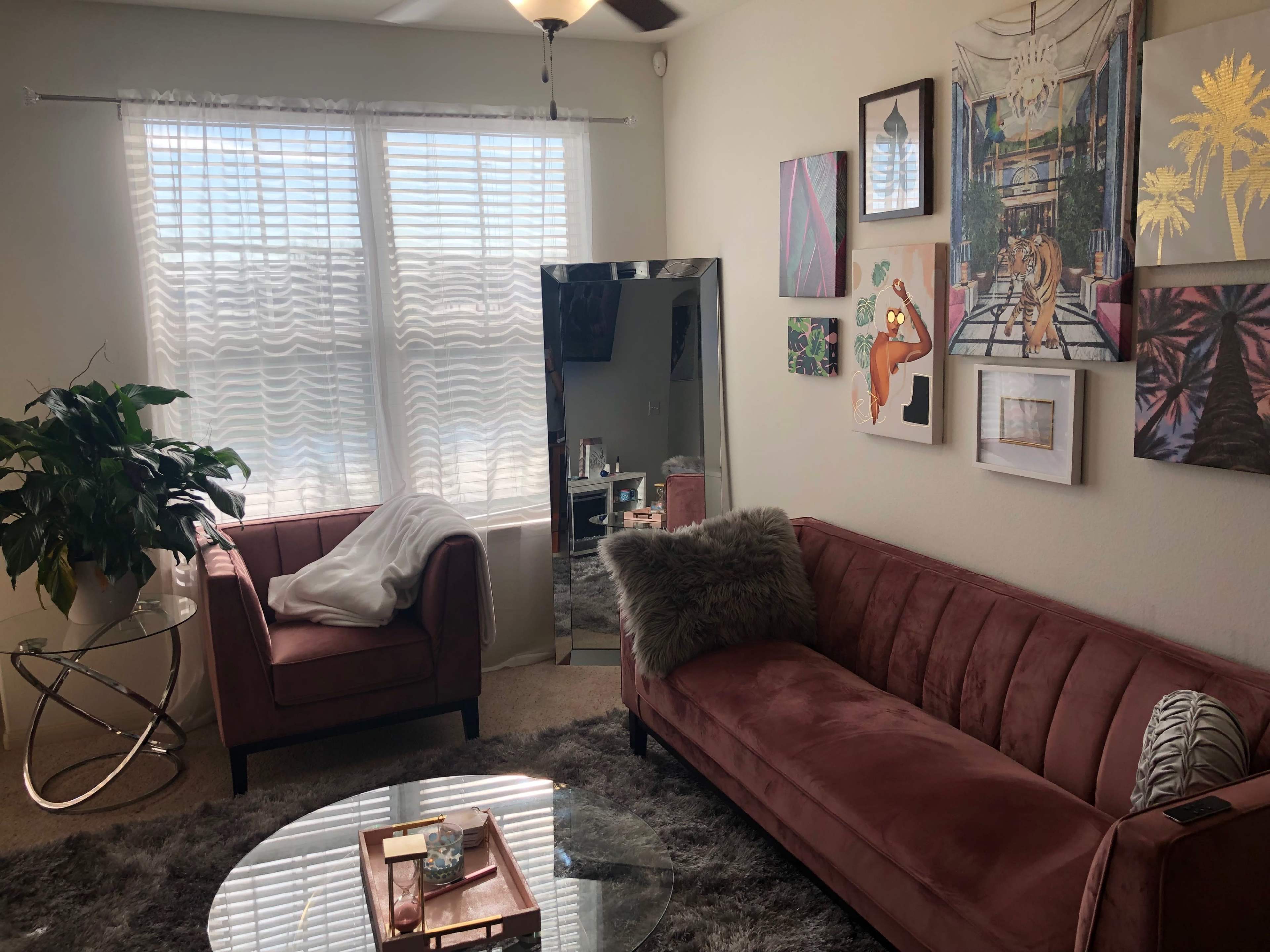 The scene features a living room with a pink velvet sofa, an armchair, a round glass coffee table, and various artworks displayed on the walls, illuminated by light from two windows.