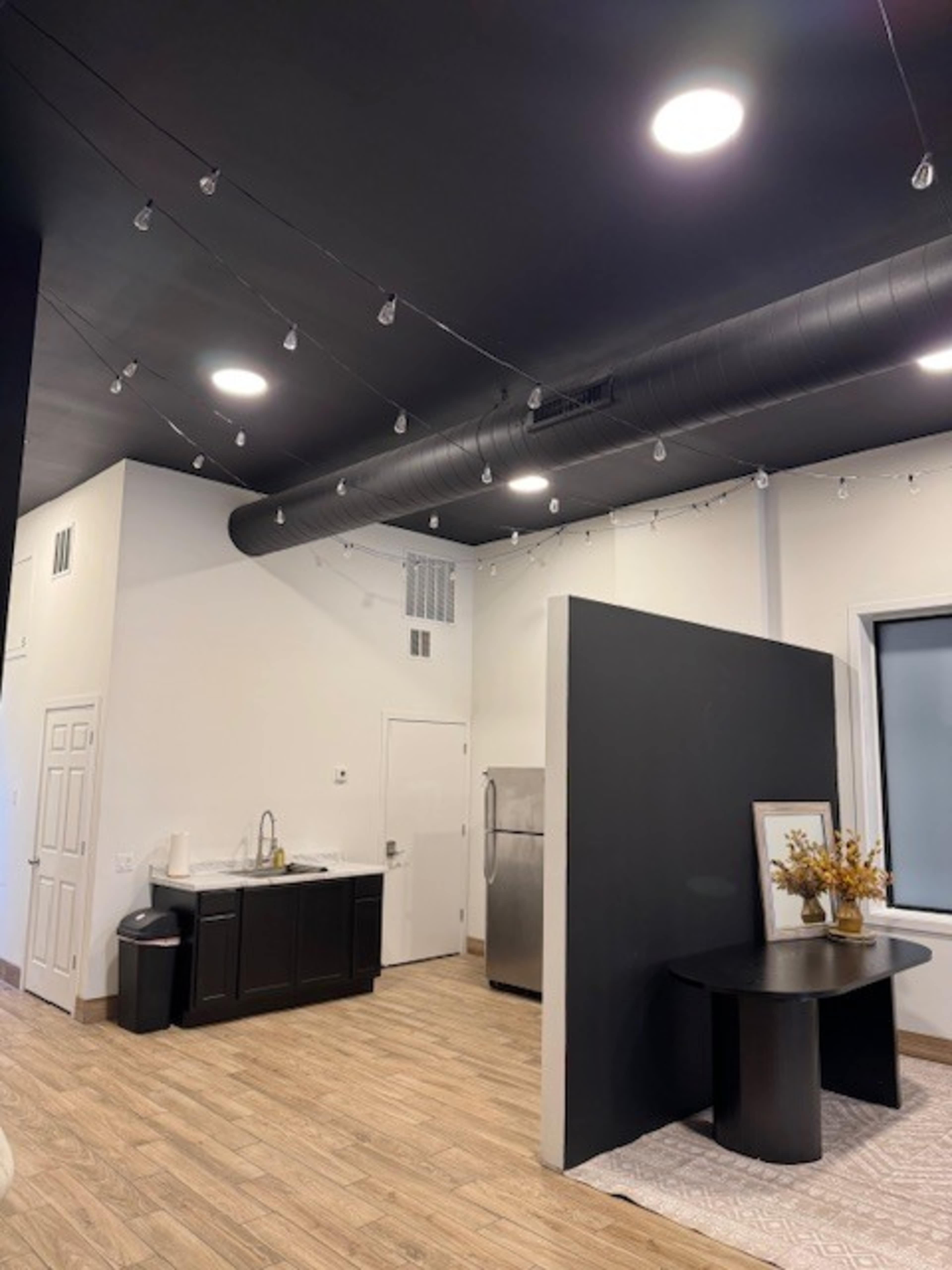 The image shows a modern, open space featuring a kitchenette with black cabinetry, a stainless steel refrigerator, and a small dining table under a black ceiling with pendant lights.