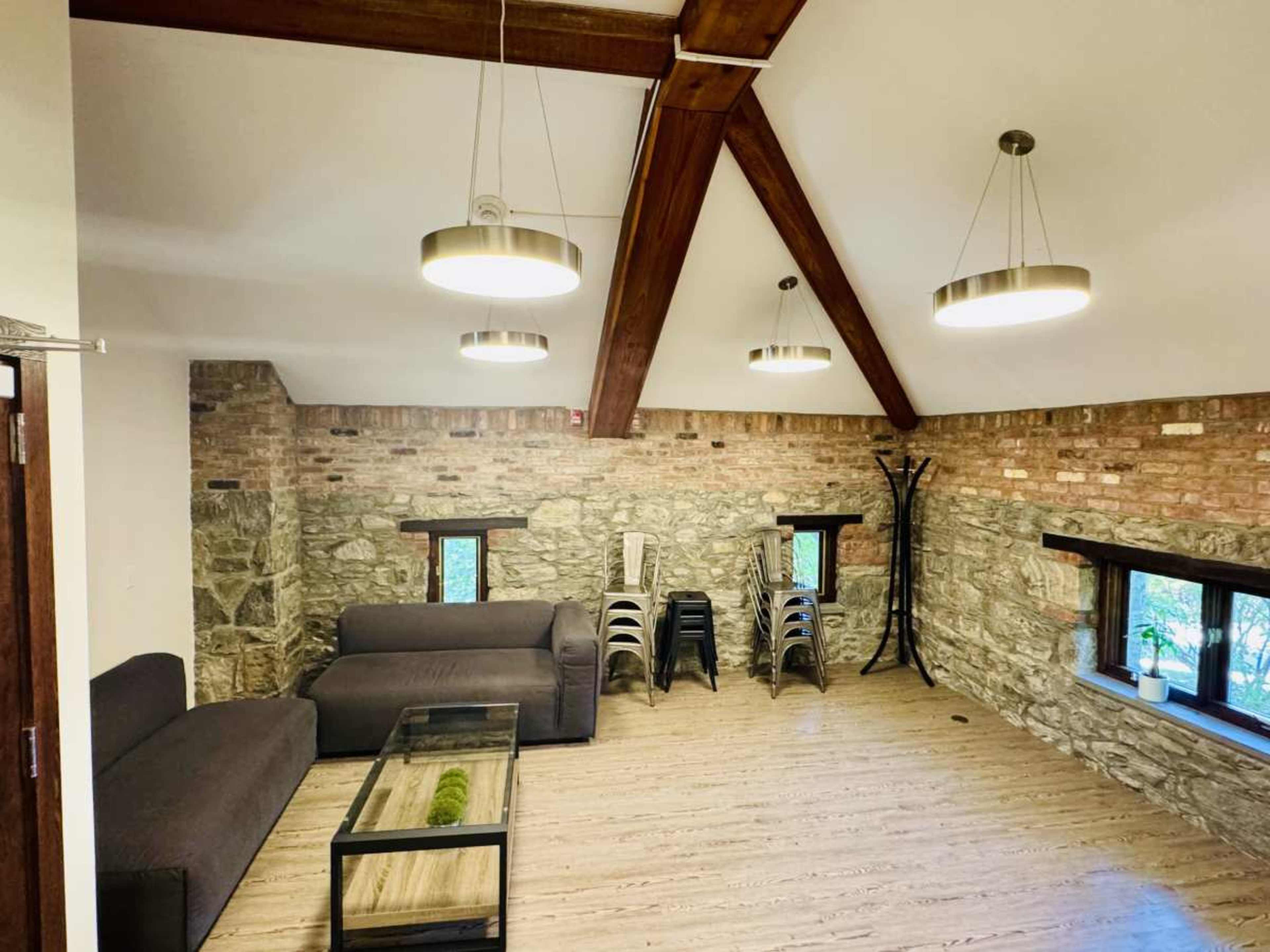 The image shows a spacious interior room with a mix of exposed stone and wooden beams, featuring a dark sofa, a glass coffee table, and stackable chairs positioned against the wall.
