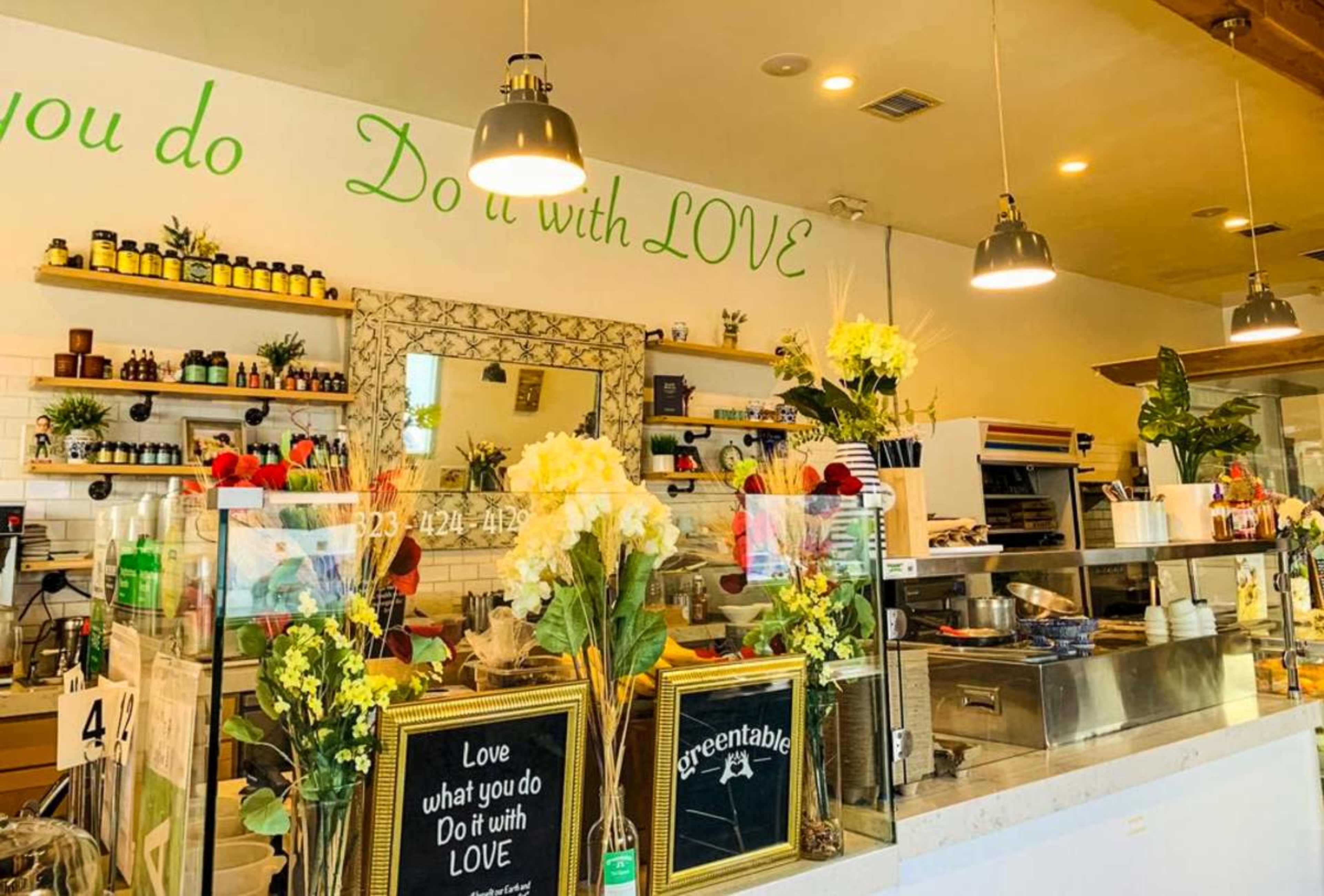The image shows a bright café interior with shelves filled with various products, floral decorations on the counter, and a wall displaying the phrase "Do it with LOVE."