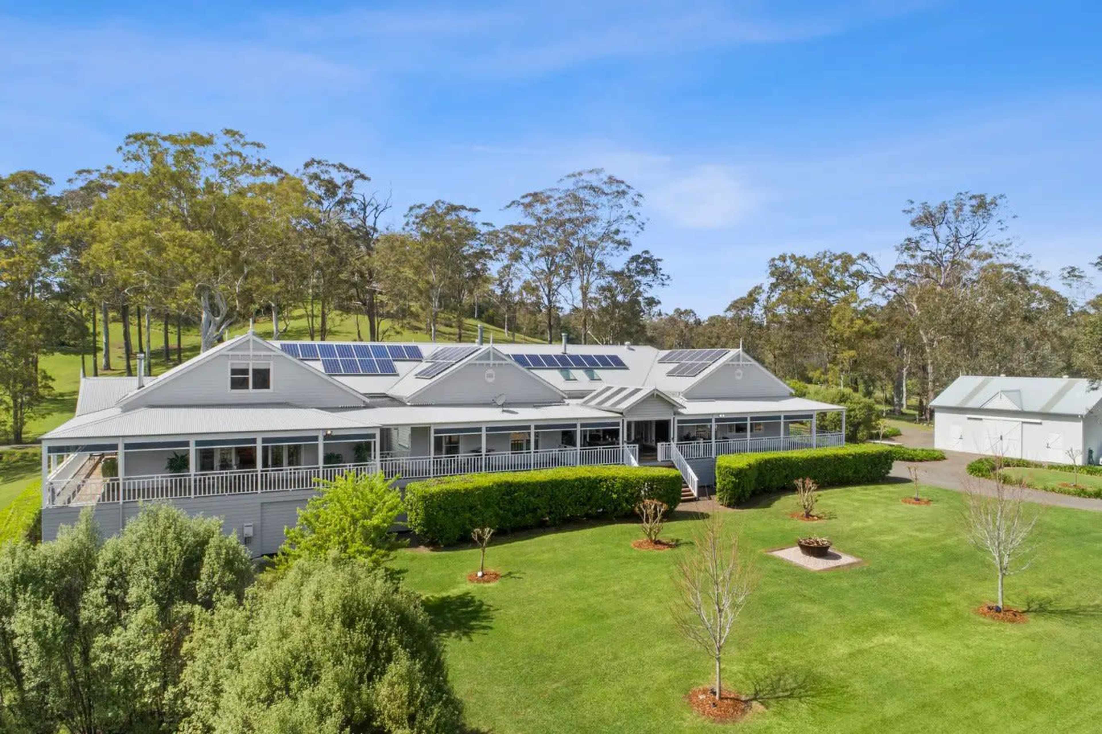 A large, modern house with a wraparound porch and solar panels on the roof is surrounded by well-maintained lawns and trees.