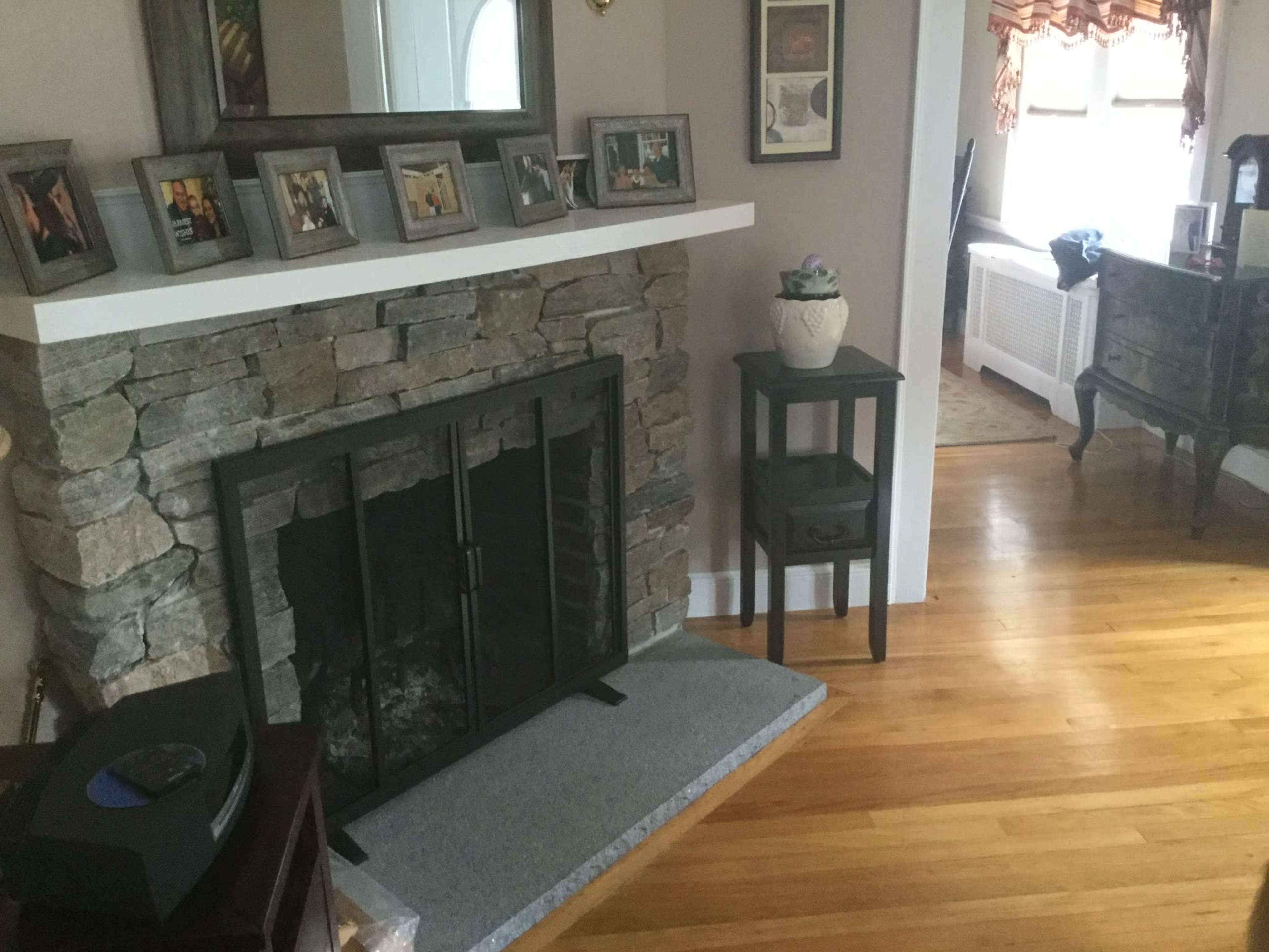 A living room with a stone fireplace, a wooden floor, framed pictures on the mantel, and a small table beside the fireplace.
