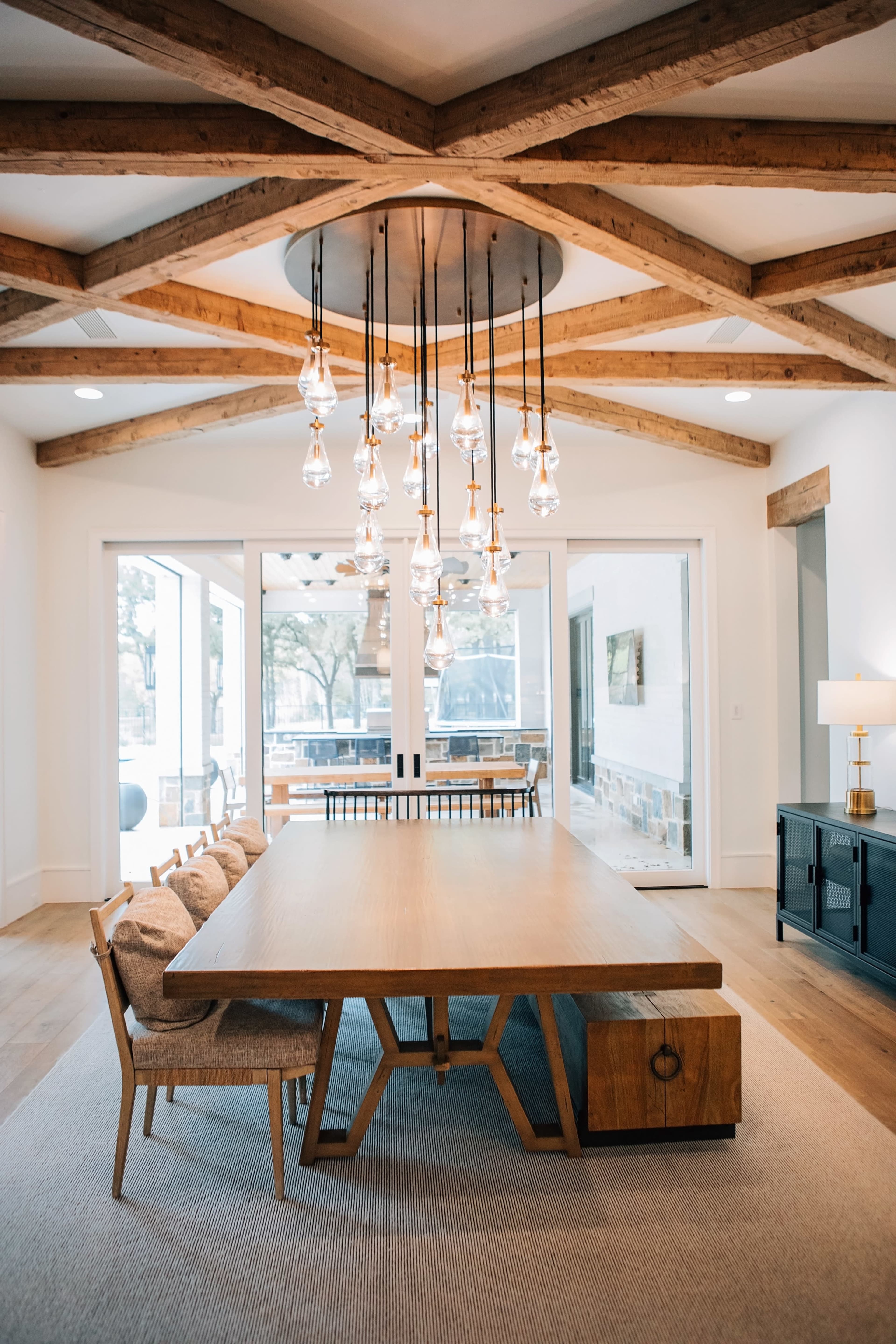 A modern dining area with a large wooden table, several upholstered chairs, and a cluster of pendant lights hanging from a wooden beam ceiling.