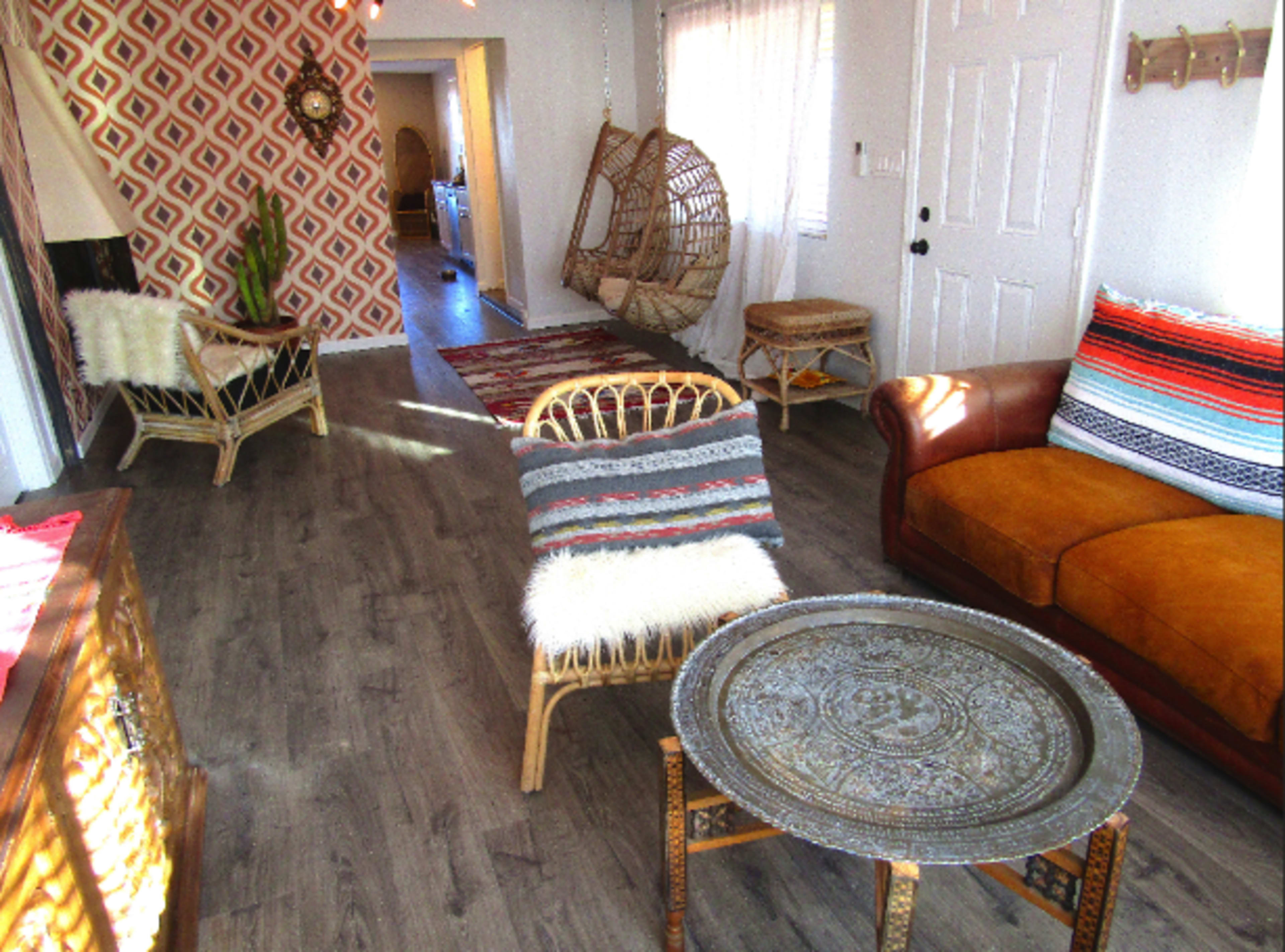 The image shows a cozy living space with a mixture of rattan furniture, a brown couch, and patterned walls, illuminated by natural light.