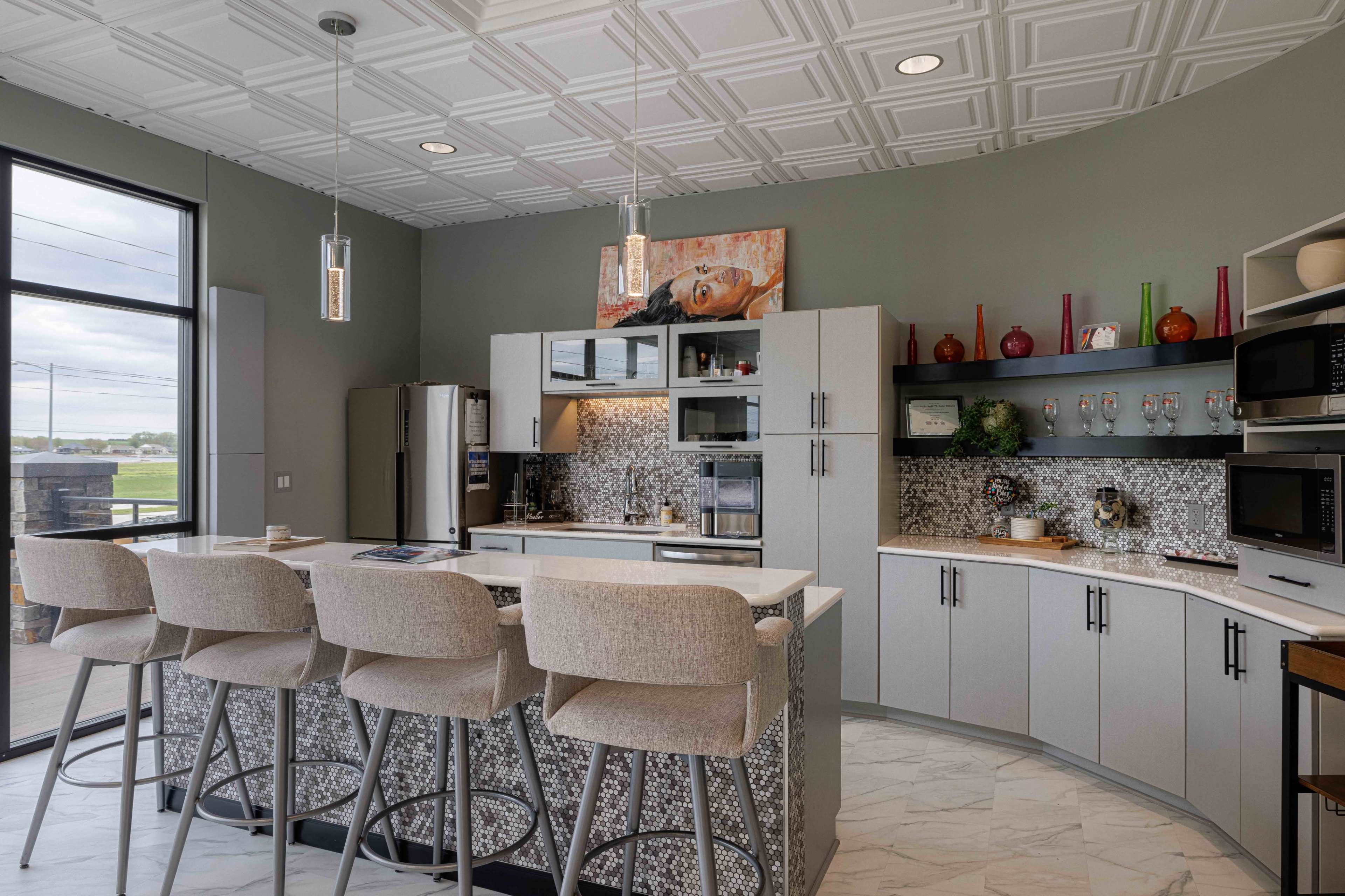 The image shows a modern kitchen with a central island, bar stools, and a mix of cabinetry and appliances, complemented by a decorative backsplash.