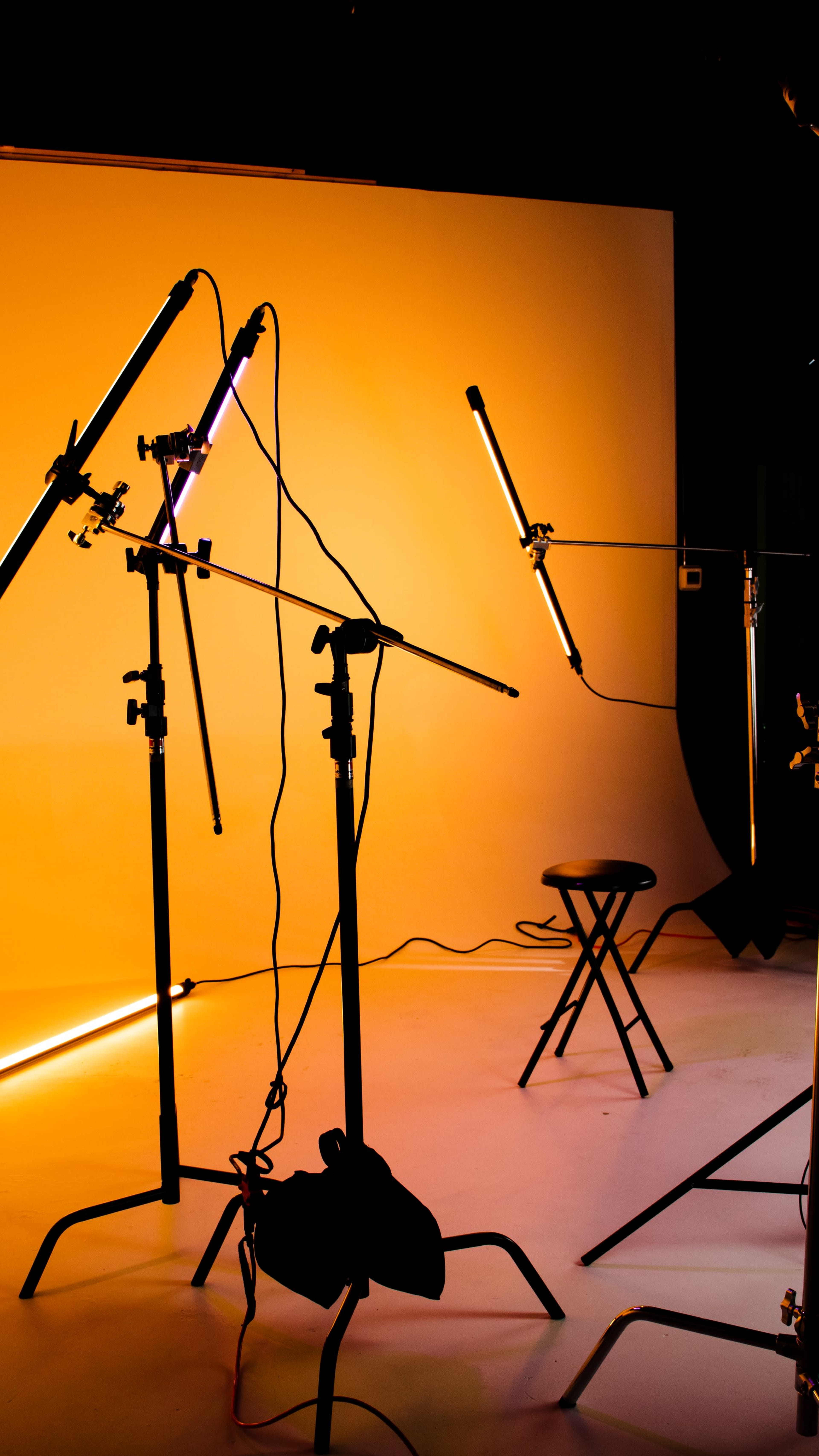 A photography studio setup features several lighting fixtures on stands directed towards a black stool against a gradient orange backdrop.