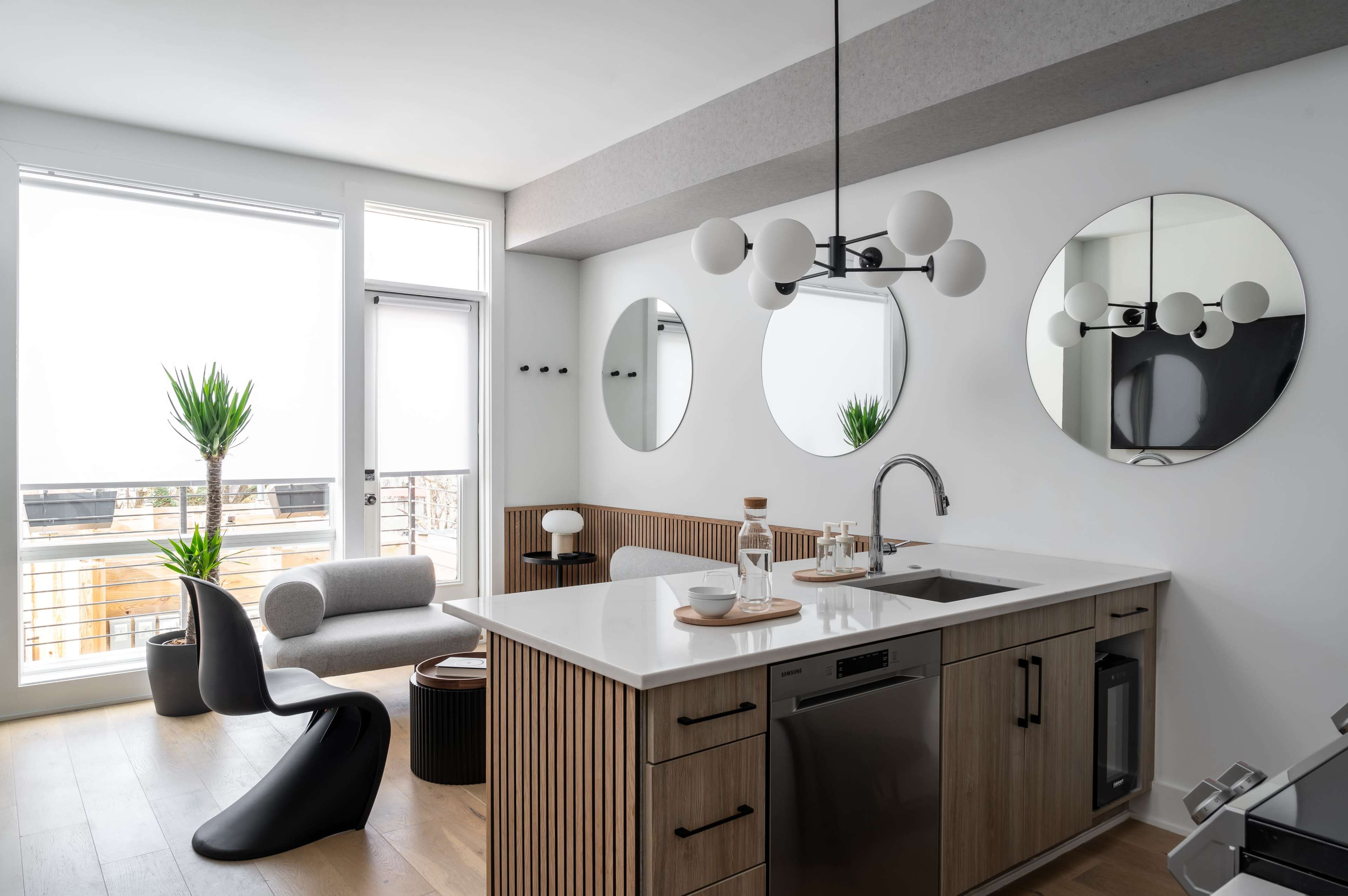 A modern kitchen with a white countertop, wooden cabinetry, and circular mirrors, complemented by plants and contemporary furniture.