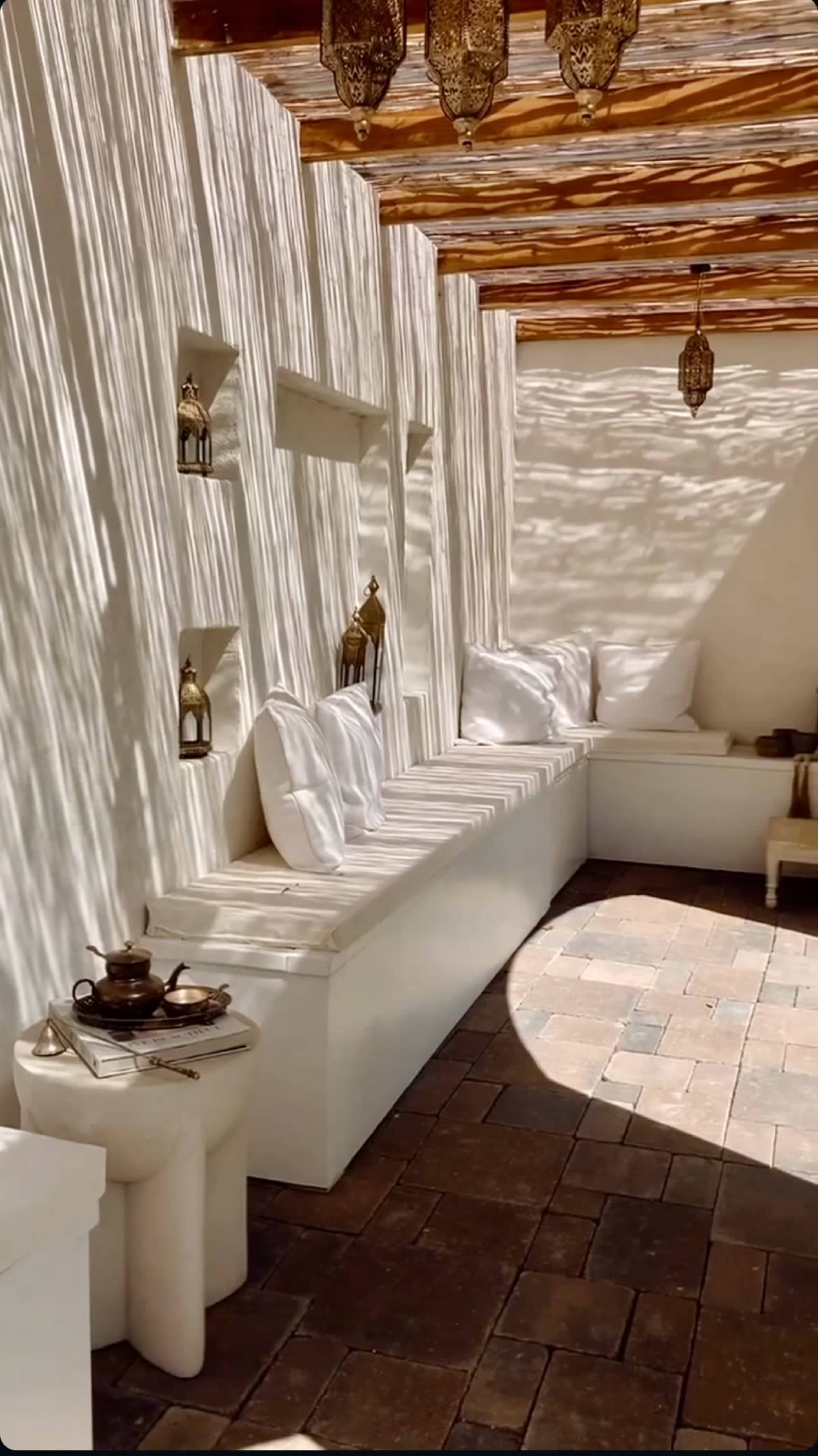 The image shows a well-lit, minimalist interior space with white walls, a long bench with cushions, and decorative lanterns casting shadows.