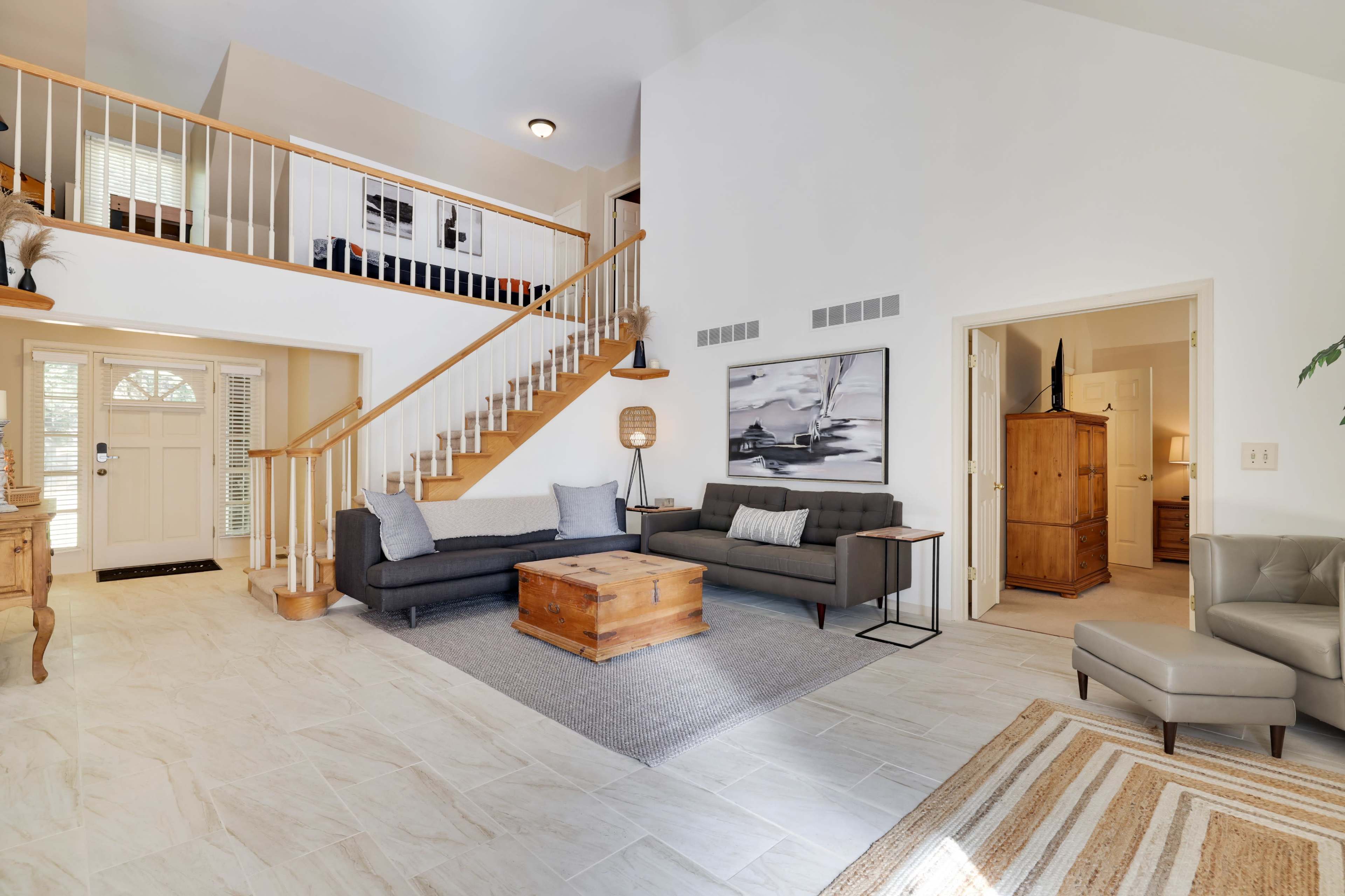 The image shows a spacious living room with a gray sofa, a black sofa, a wooden coffee table, and a staircase leading to an upper level.