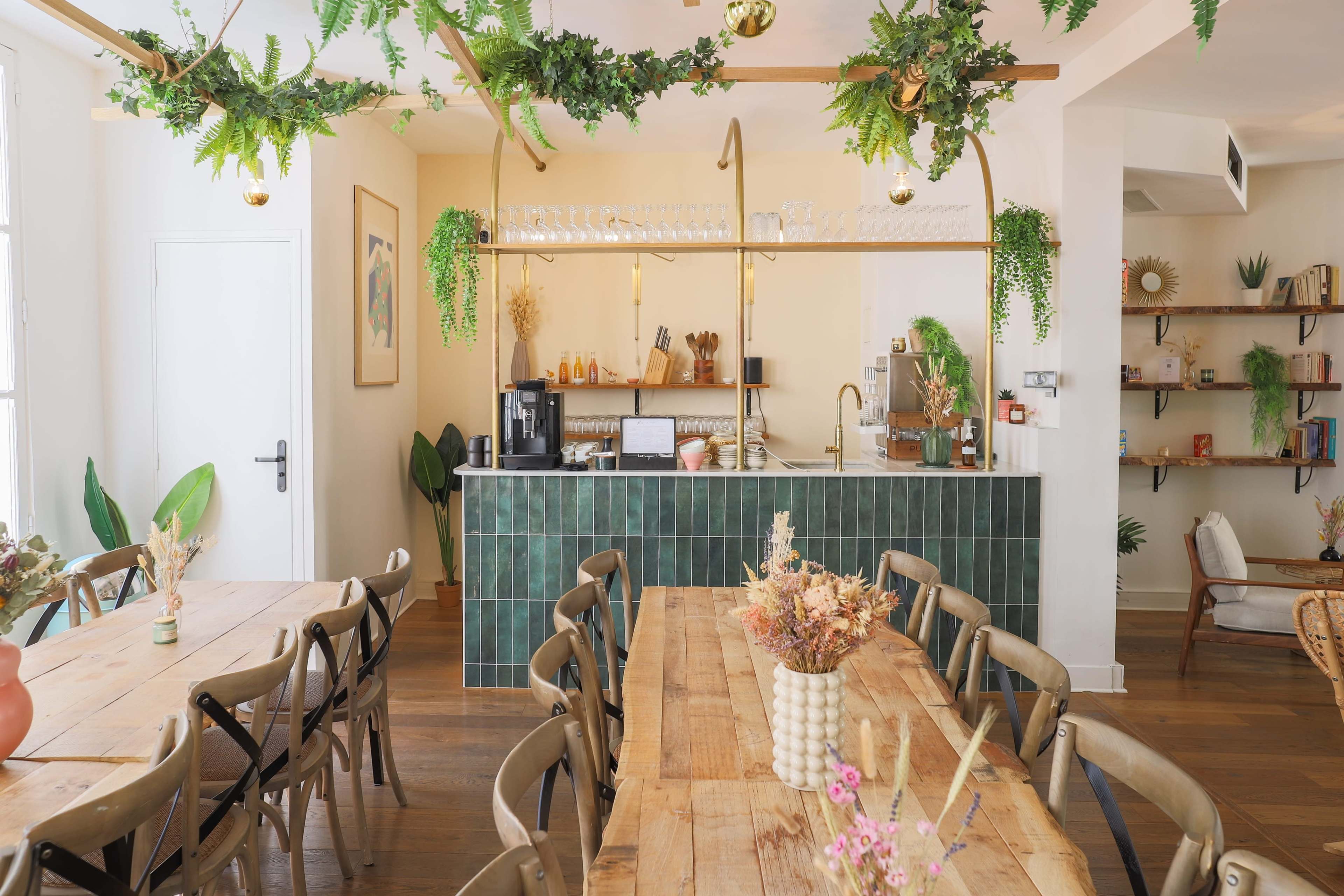 The image shows a bright, modern cafe with wooden tables, green tile accents, and a bar area adorned with plants and glassware.