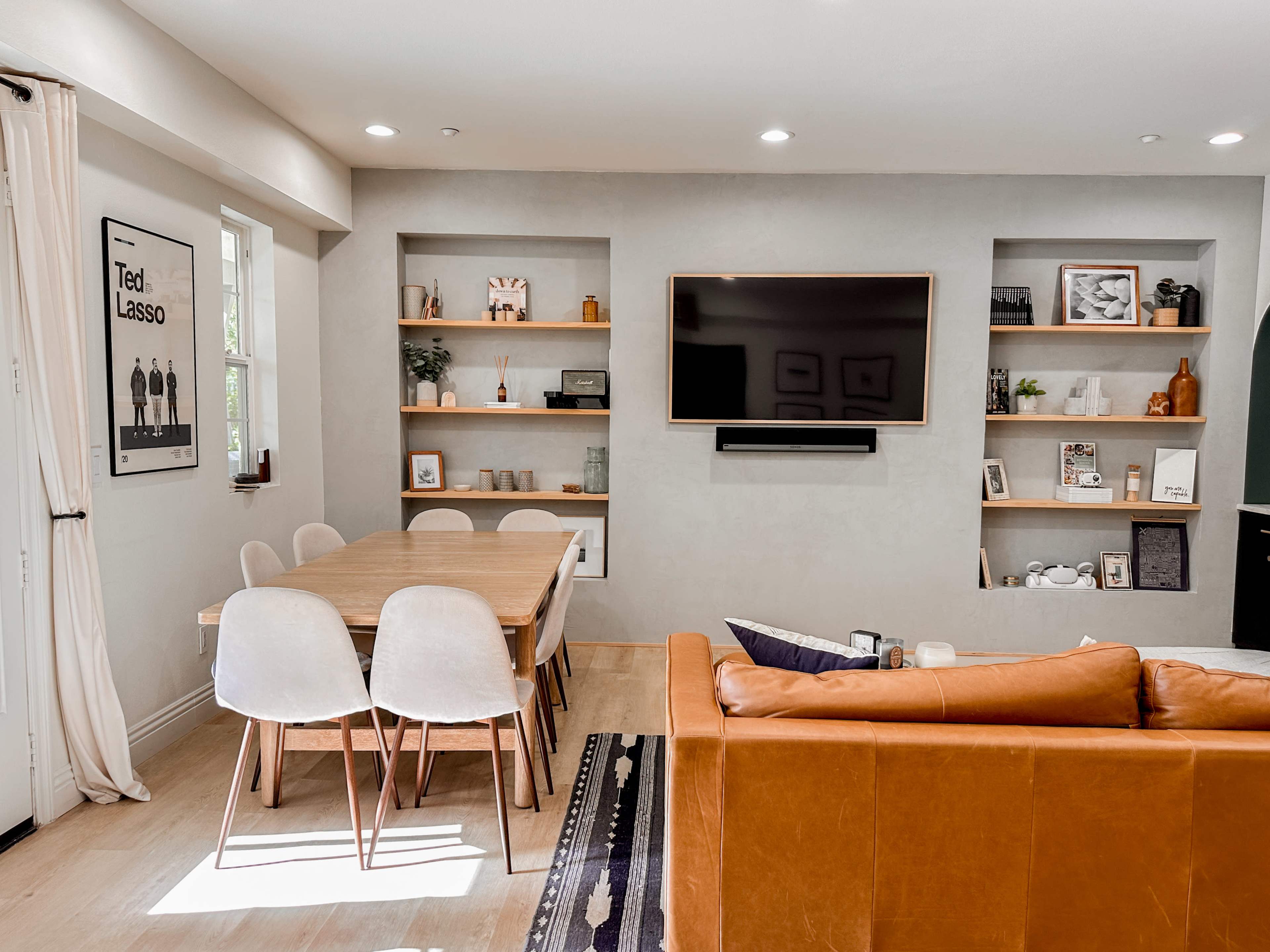 The scene shows a modern living room with a brown leather sofa, a wooden dining table surrounded by white chairs, and a wall-mounted television above floating shelves filled with decorative items.