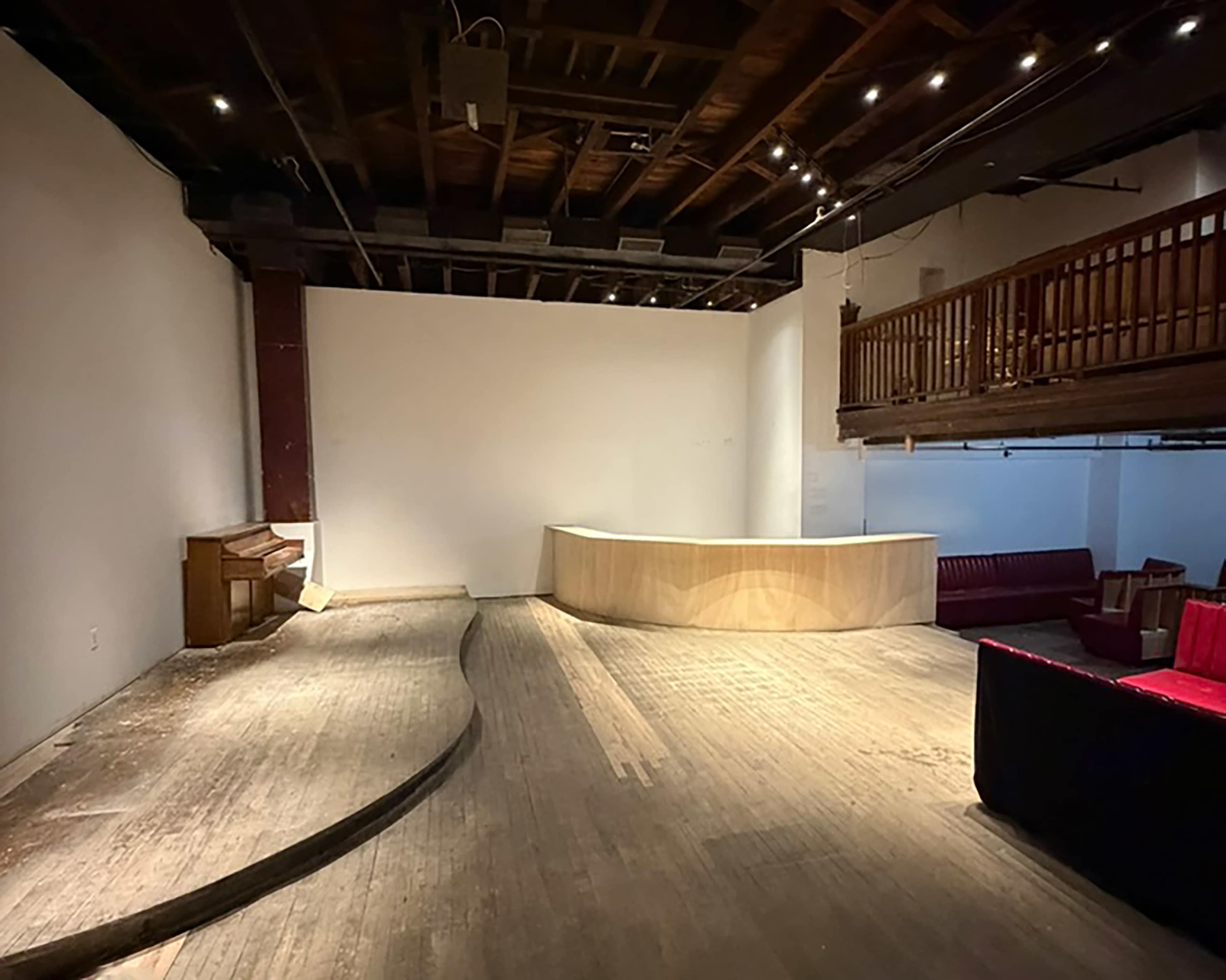 The image shows an empty indoor space featuring a wooden floor, a curved wooden reception desk, a piano against one wall, and a small elevated area with a railing.