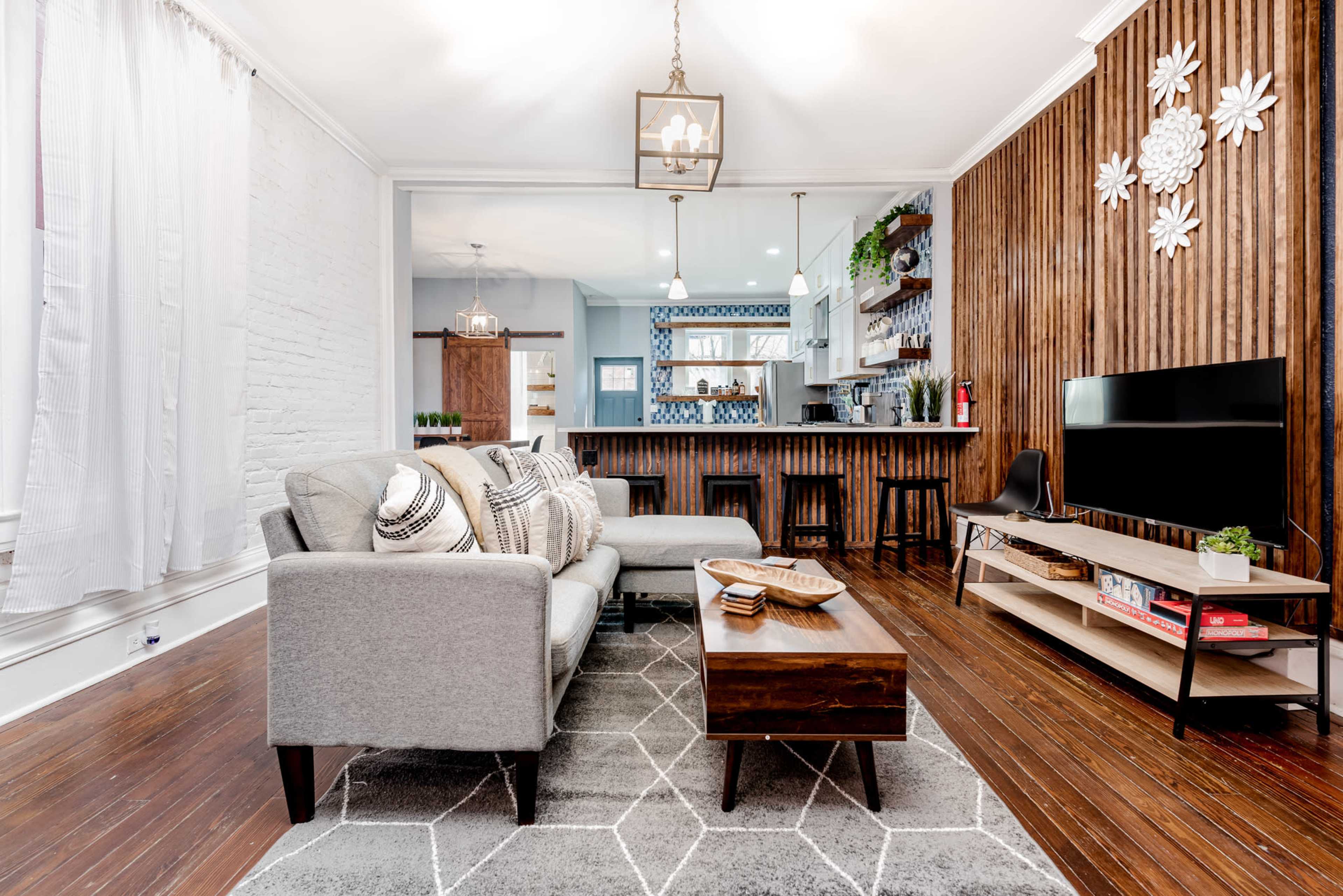 The living room features a gray sectional sofa, a wooden coffee table, and a television, with a backdrop of wood-paneled walls and a kitchen visible in the background.