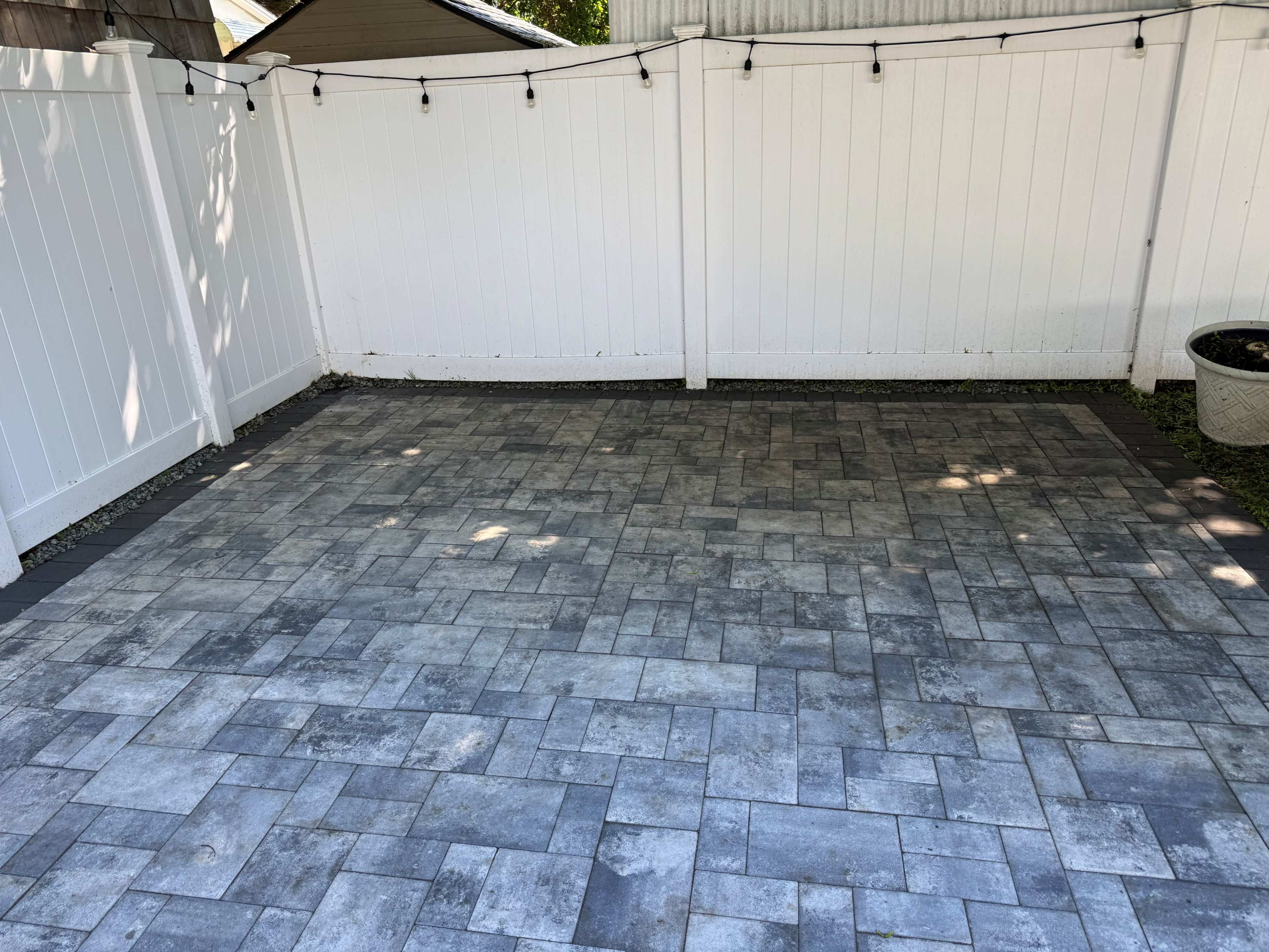The image shows a paved patio area with gray interlocking stones, surrounded by a white fence and illuminated by string lights above.
