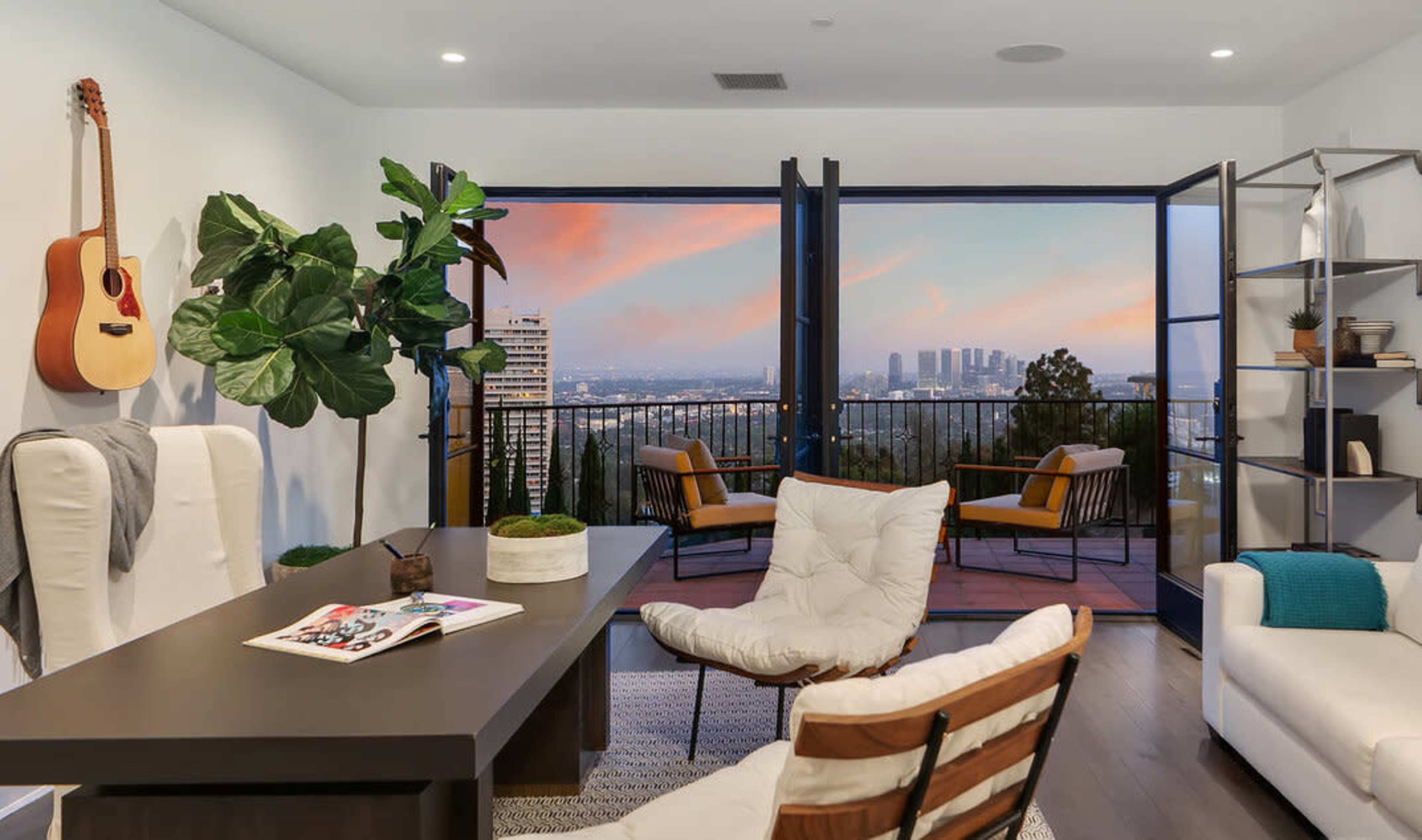 A modern living space with a desk and seating, large windows opening to a city view, and a plant and guitar on display.