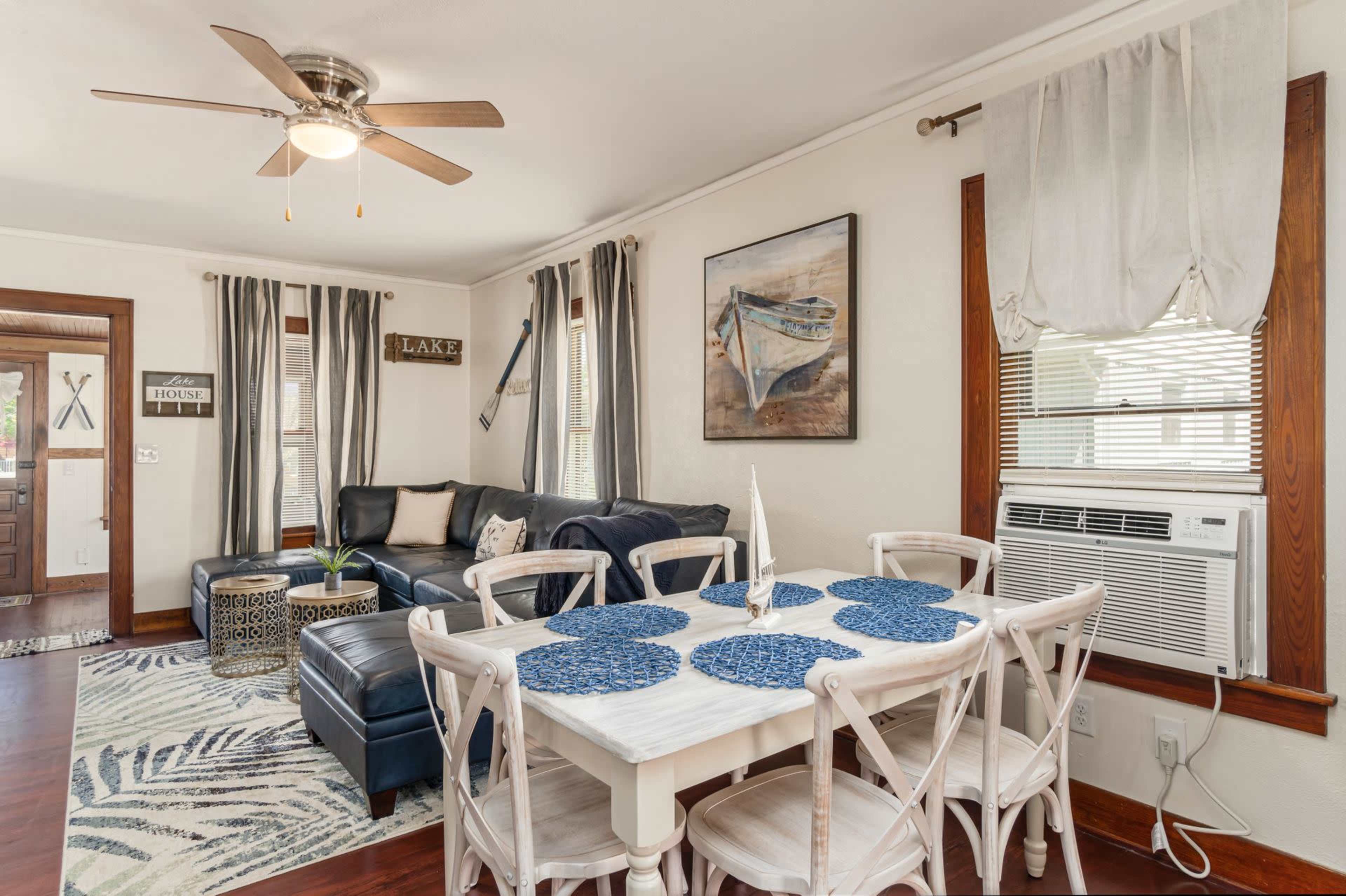 A living and dining area features a navy sofa, a white dining table with blue placemats, and an air conditioning unit beneath a window.