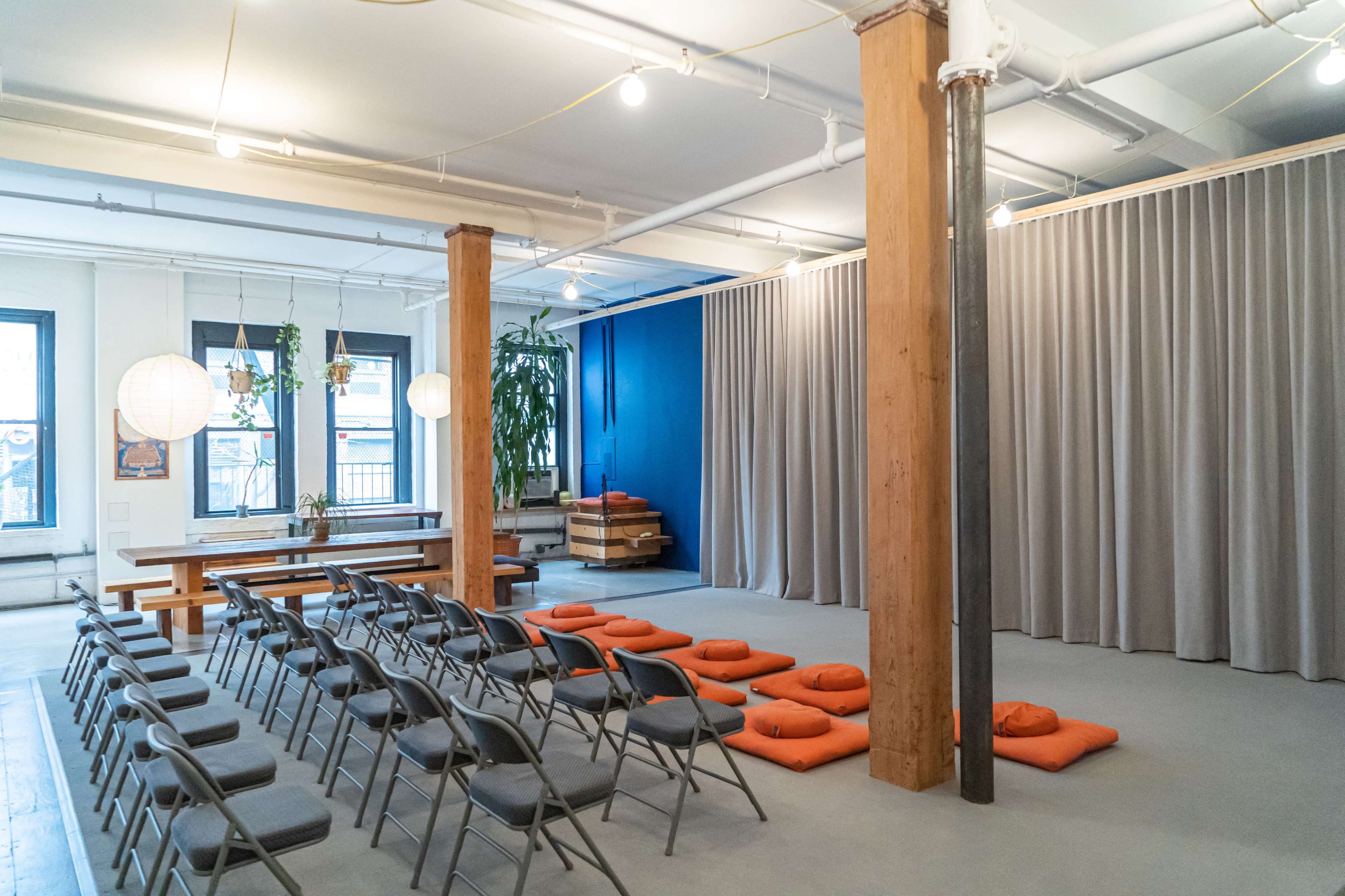 A spacious room with rows of folding chairs facing a curtain, orange cushions on the floor, and plants in the windows.