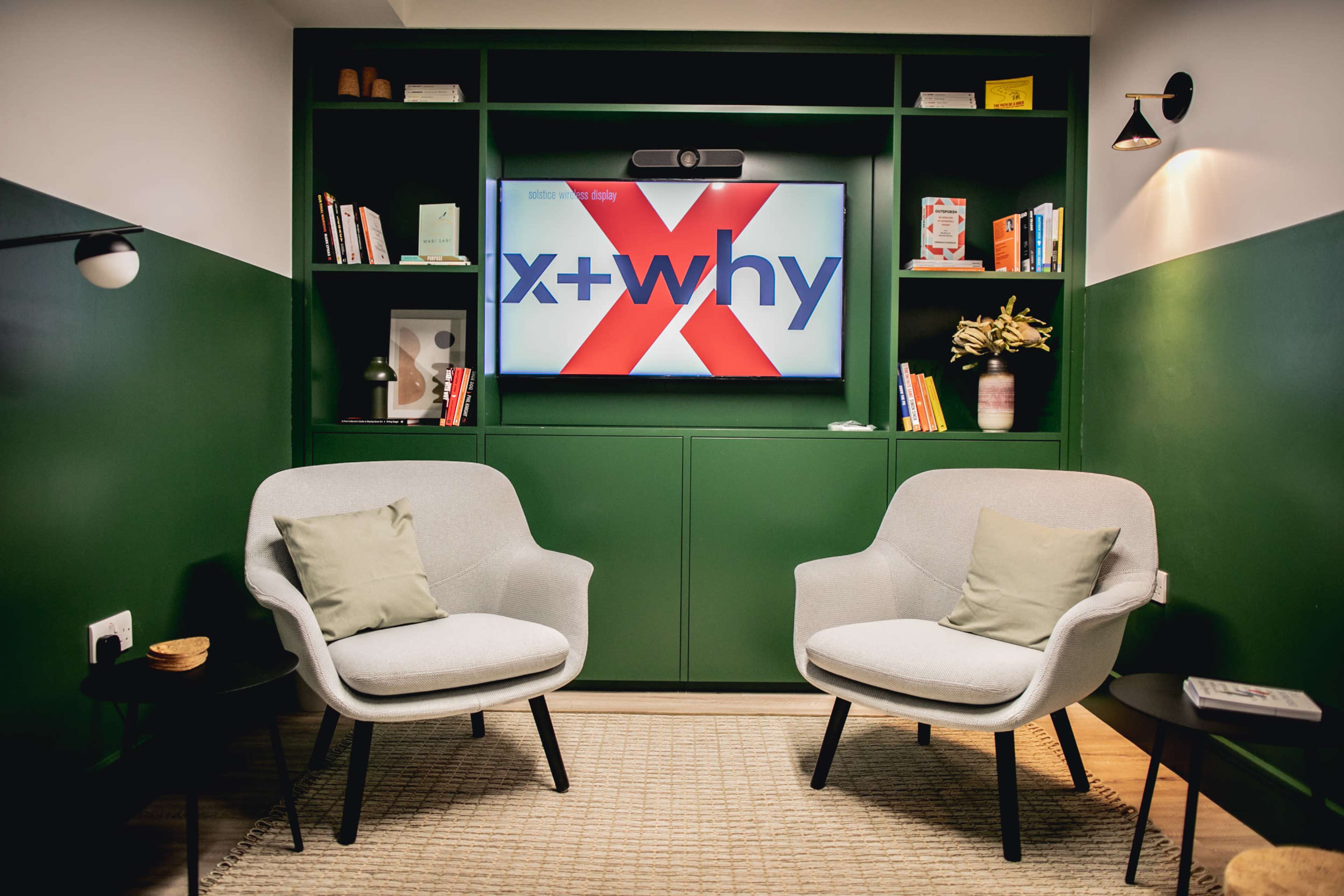 A cozy seating area features two light gray chairs facing a green wall with a TV screen displaying "x+why" and shelves filled with books and decorative items.