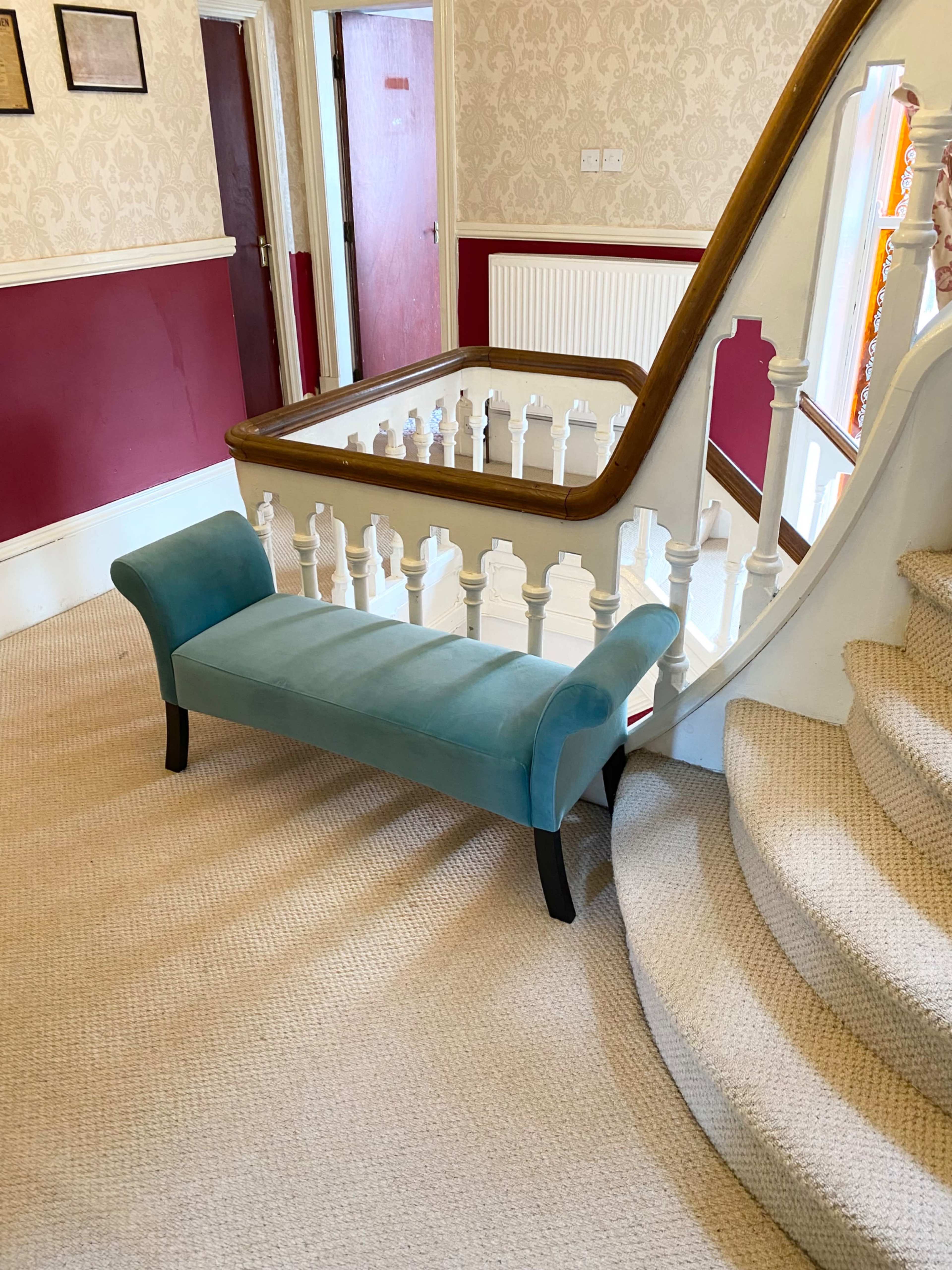 A light blue bench is positioned near the base of a staircase in a hallway with patterned wallpaper and red accents.