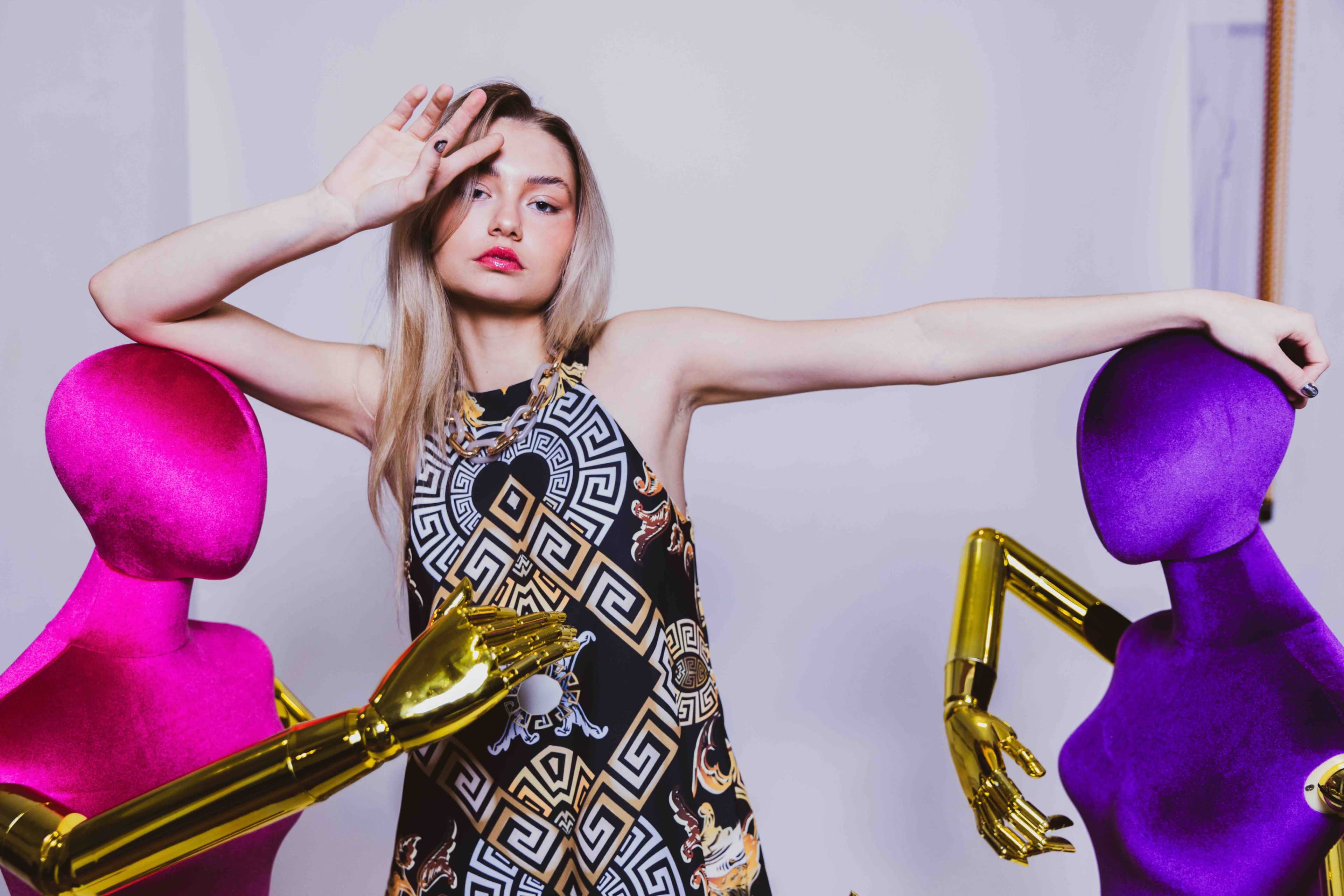 A model poses between two colorful mannequins, one pink and one purple, while wearing a patterned dress.