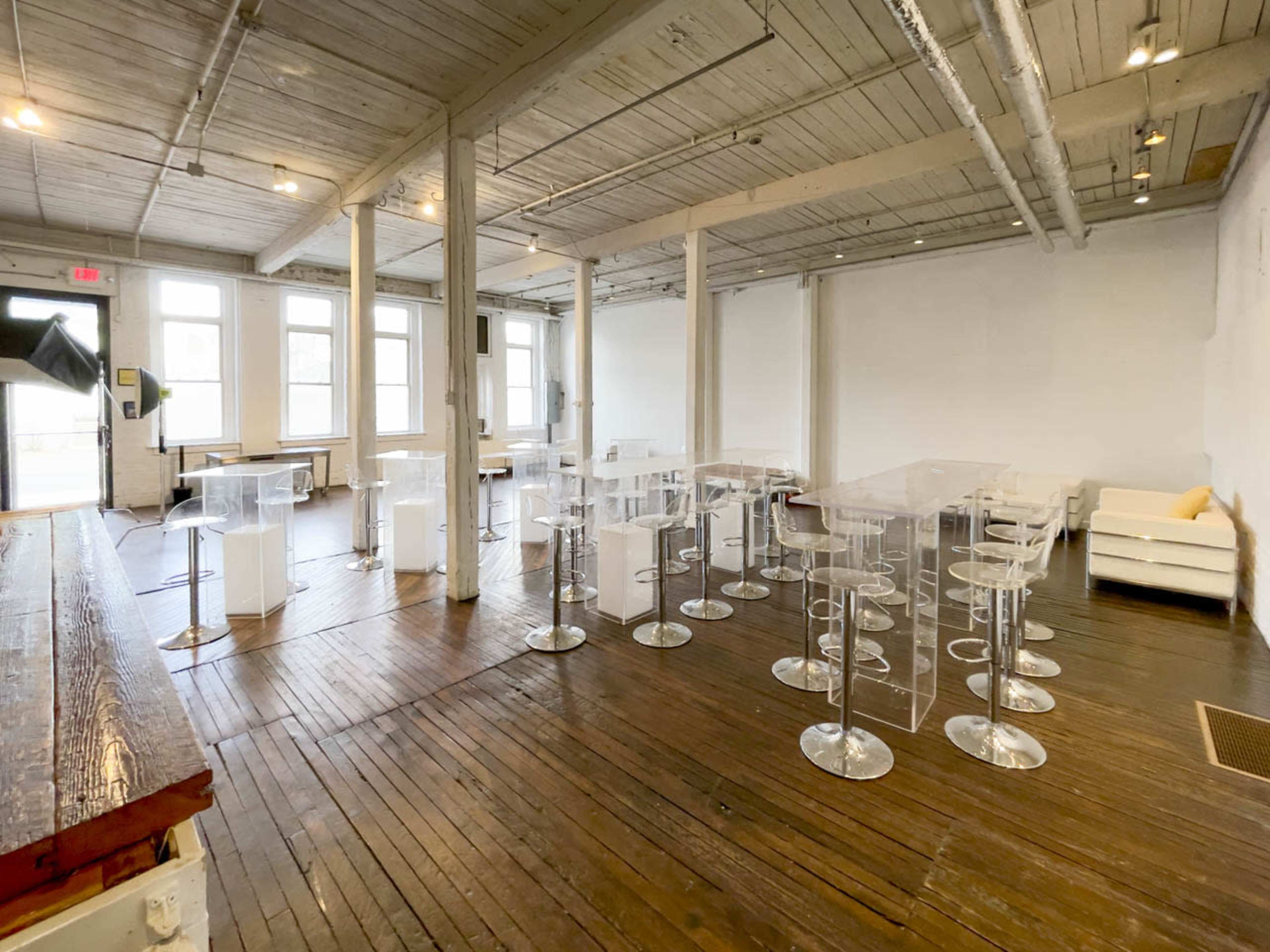 A spacious room with wooden floors, featuring transparent acrylic tables and stools arranged in a modern layout, alongside a few white seating areas.