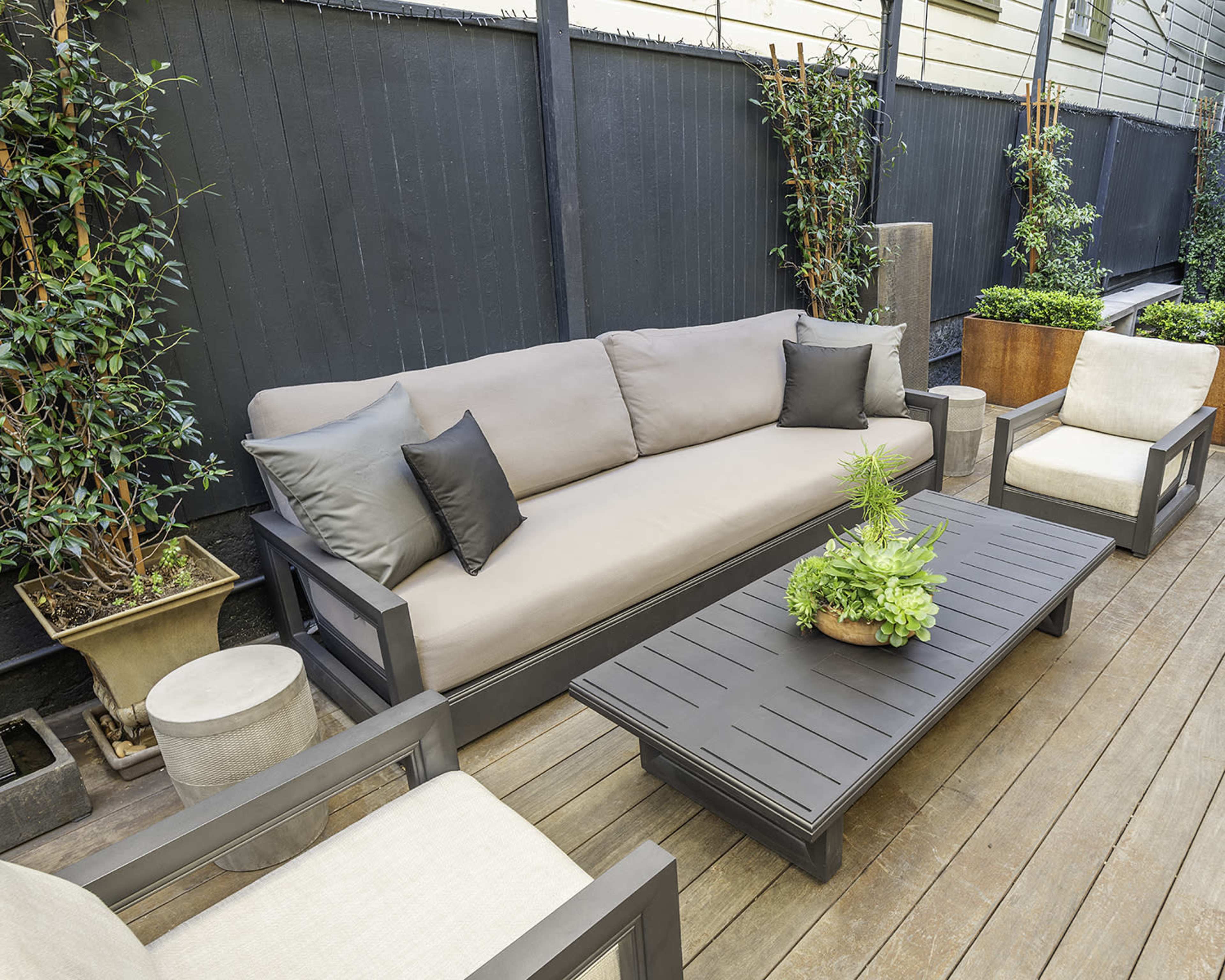 The image shows a modern outdoor seating area with a gray sectional sofa, two armchairs, and a wooden coffee table surrounded by potted plants.