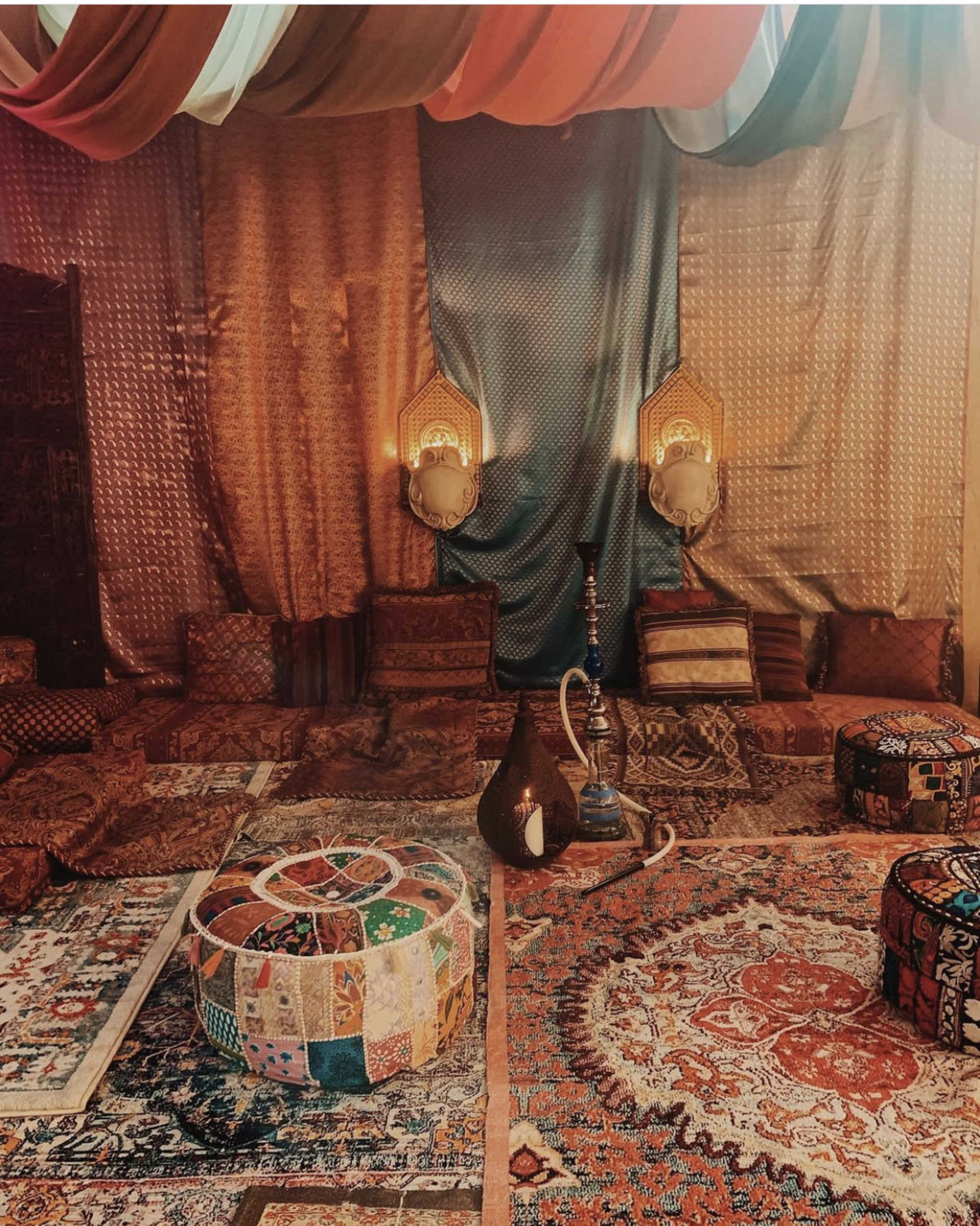 A richly decorated room with patterned rugs, cushions, and draped fabrics, featuring a hookah and a colorful pouf in the center.