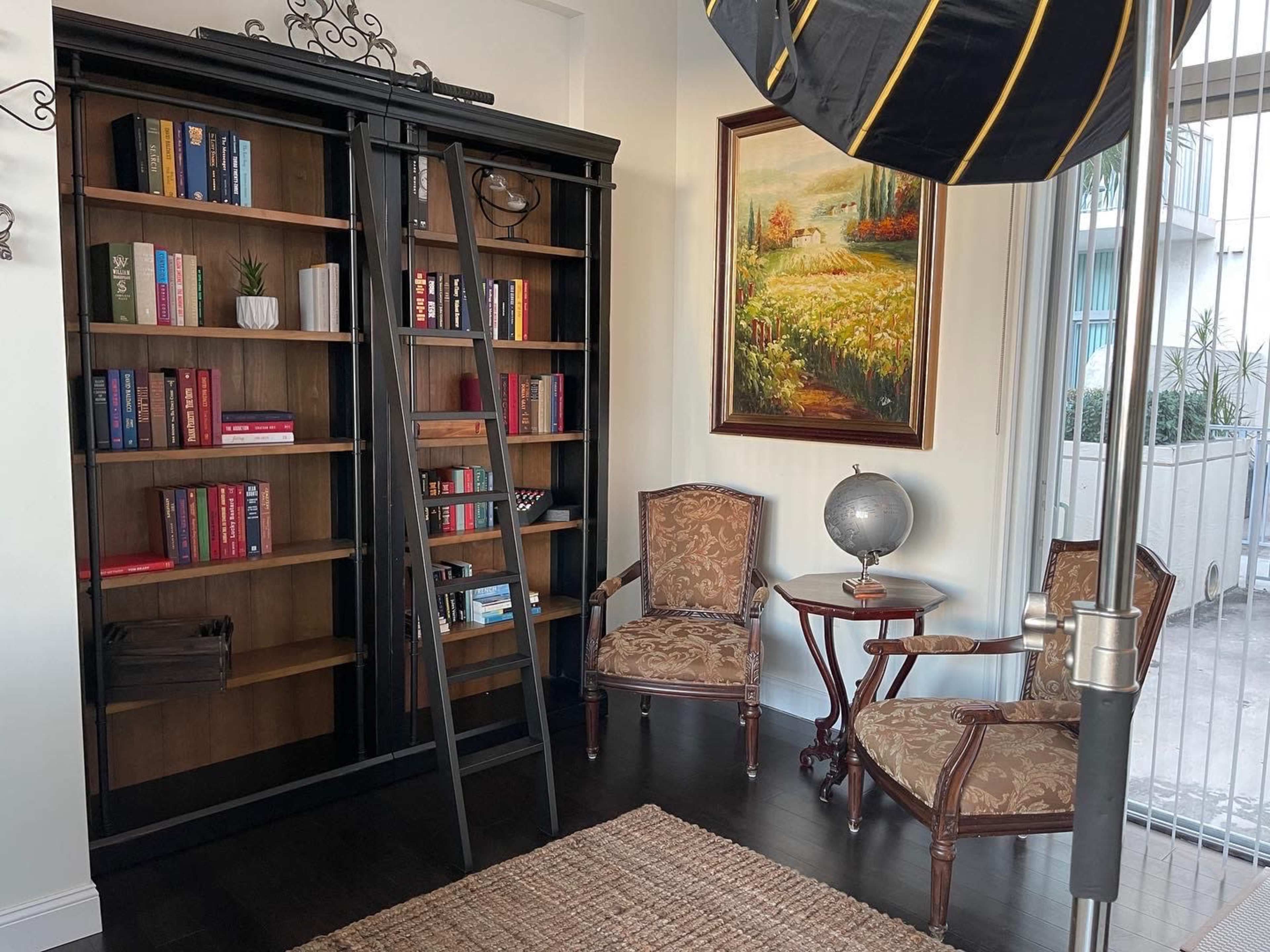 The image shows a room with a bookshelf featuring several books, a ladder leaning against it, two vintage chairs, a small table with a globe, and a large painting on the wall.