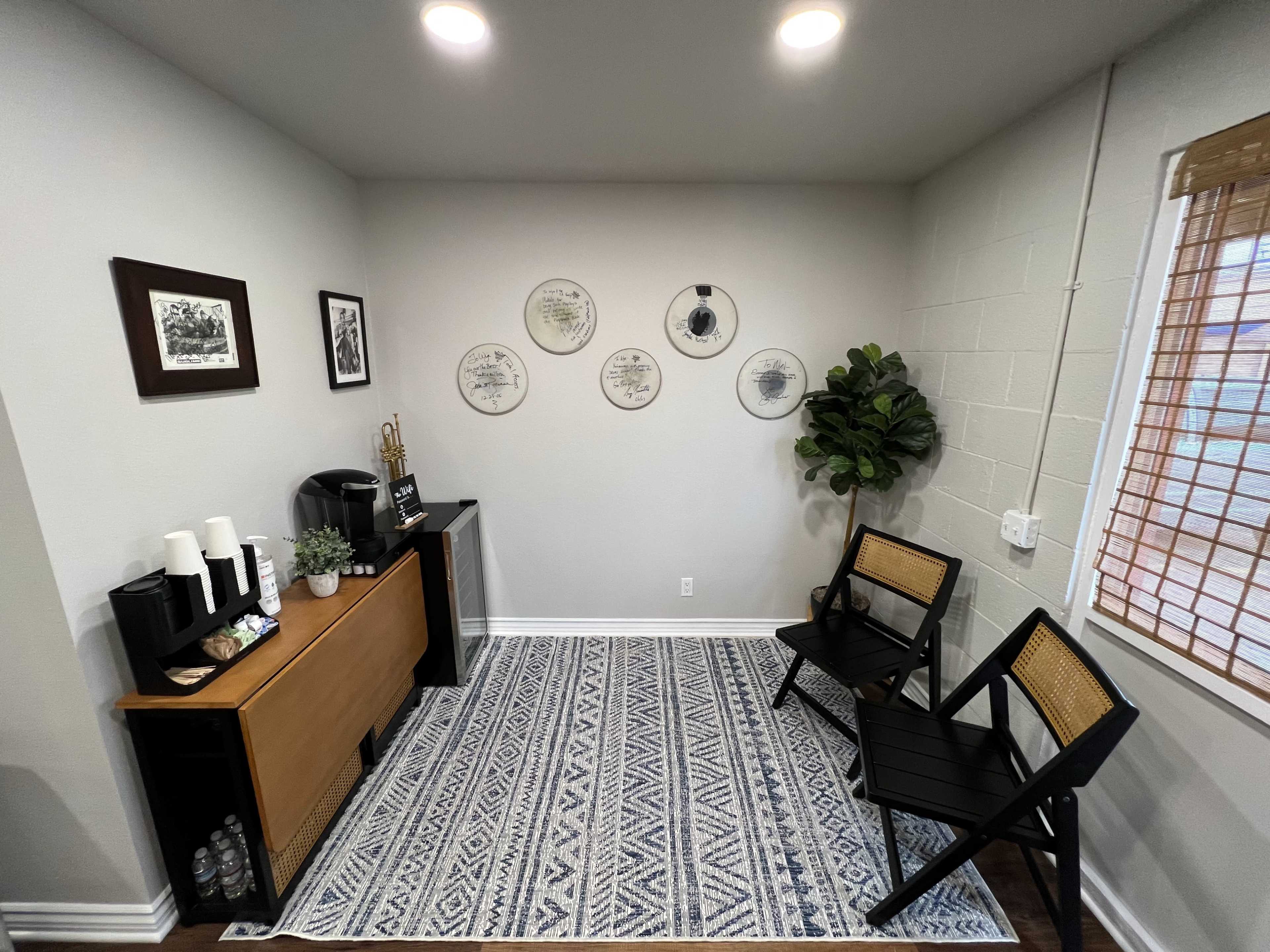 The image shows a small, well-lit room with a coffee station, two chairs, decorative wall art, and a potted plant.