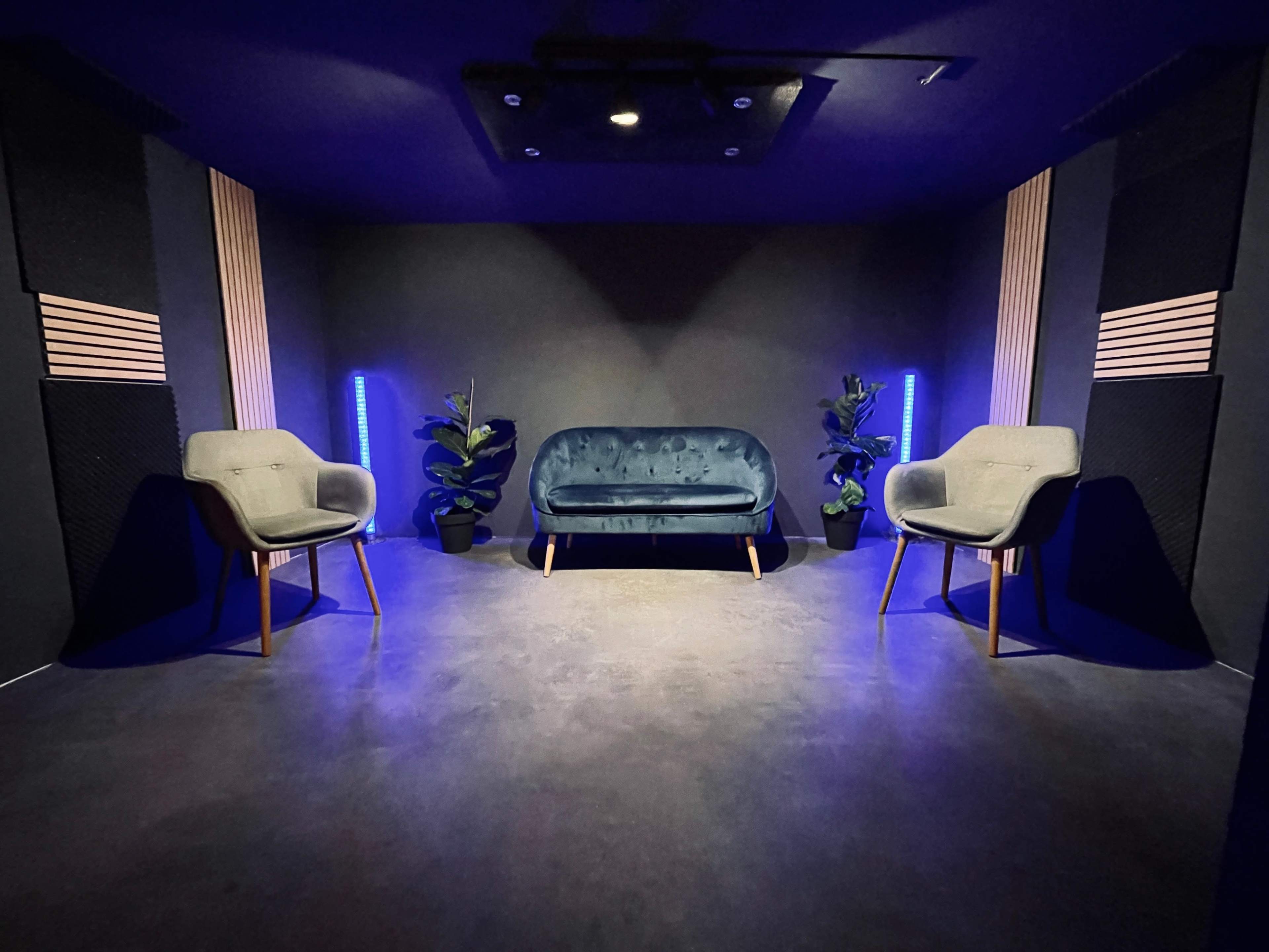 A modern room with a blue upholstered sofa and two light gray chairs, illuminated by blue lighting and flanked by potted plants.