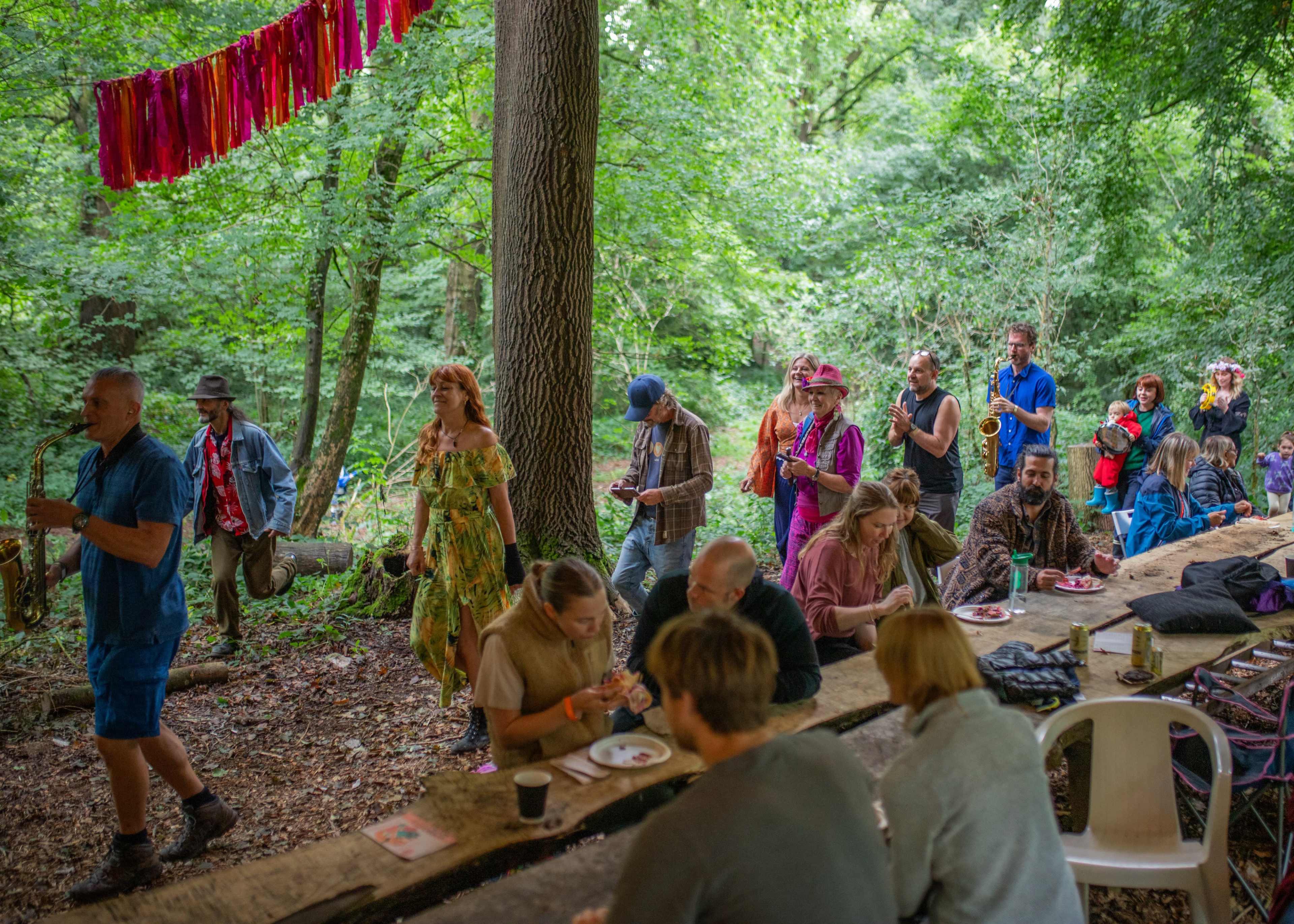 A group of people is gathered in a wooded area, walking along a table set for a meal, while others are seated and eating.