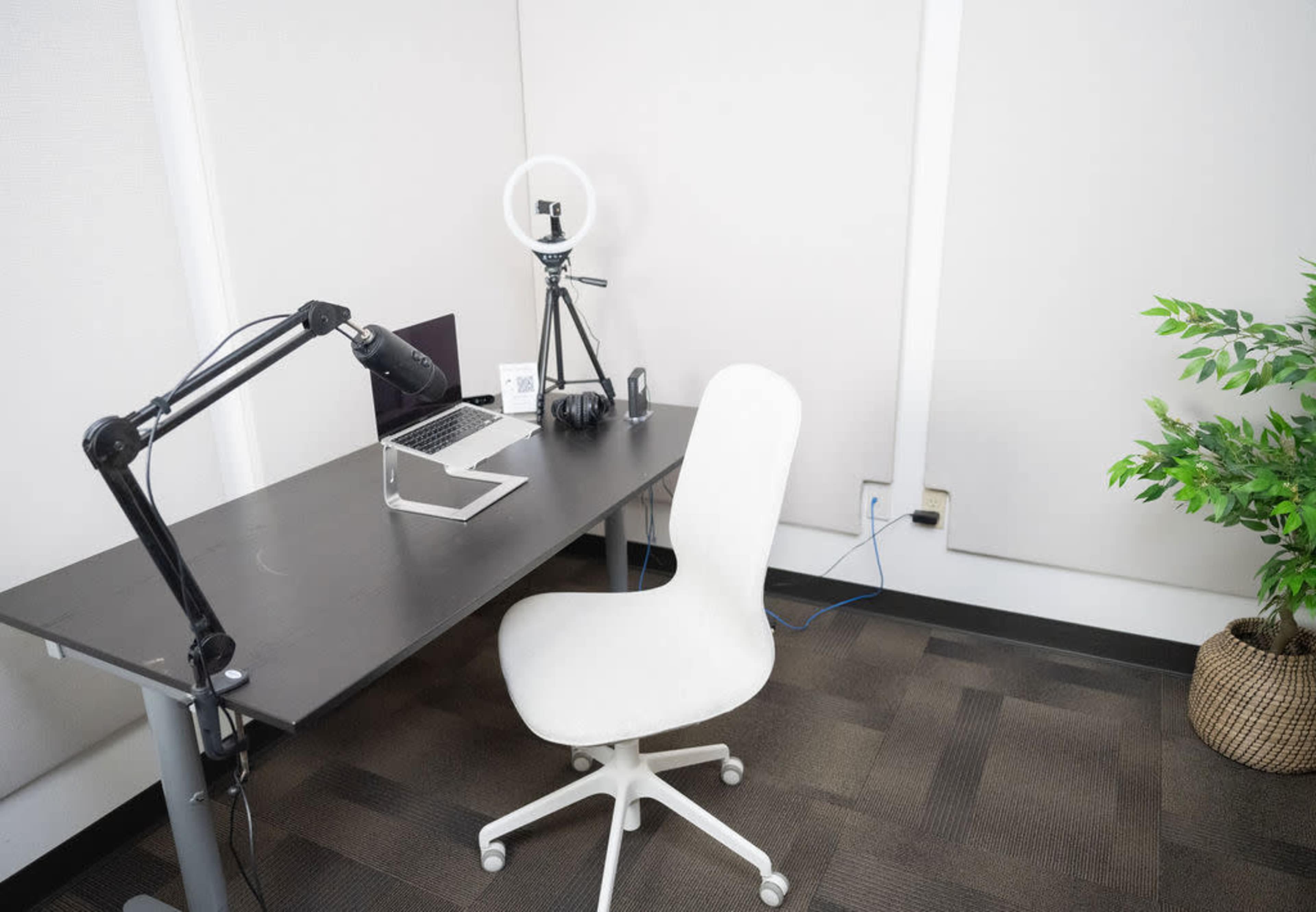 A minimalistic office space featuring a desk with a laptop, a microphone on an arm, a ring light, a white chair, and a potted plant.