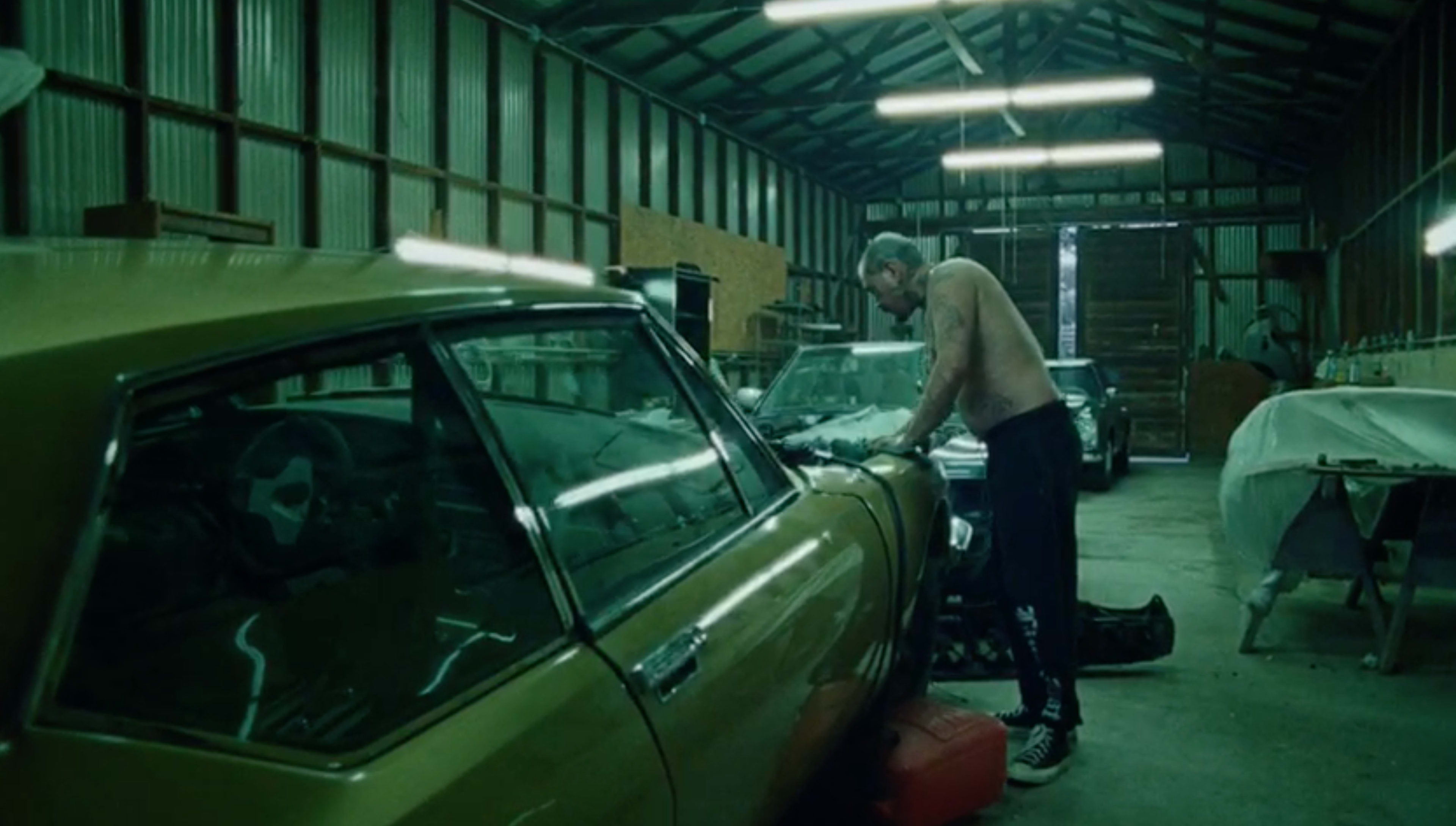 A shirtless man works on a classic car inside a dimly lit garage filled with various vehicles and tools.