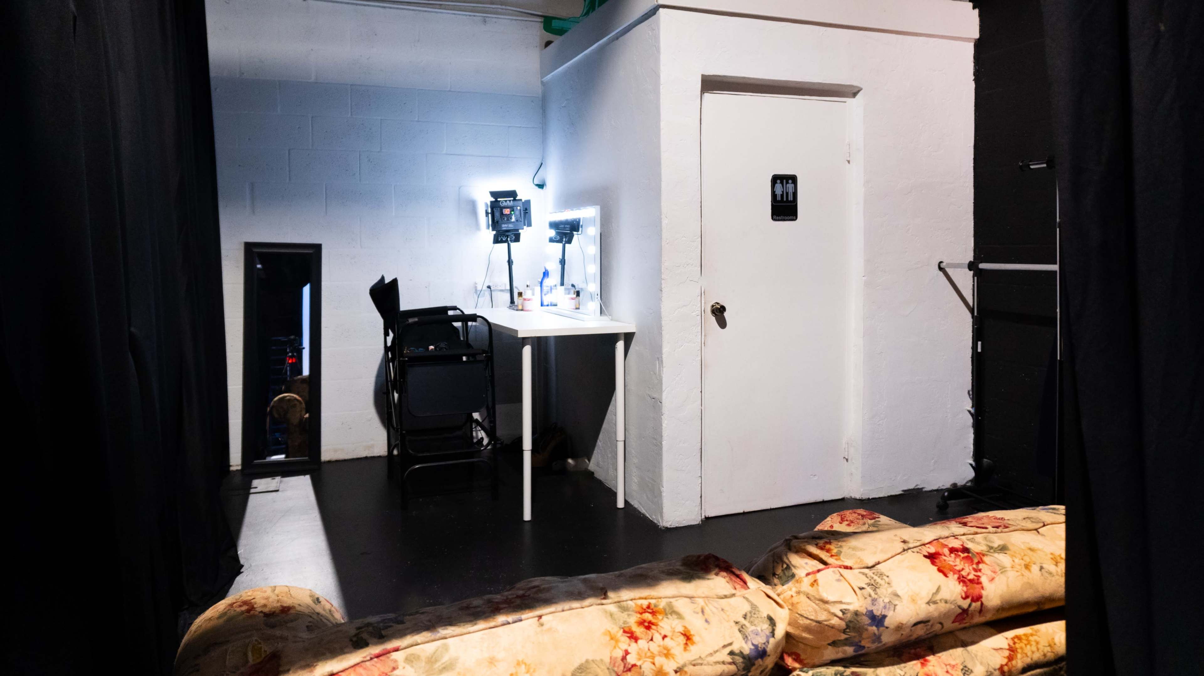 The image shows a small, dimly lit room with a table and chair set up near a white door, while a floral-patterned couch is positioned in the foreground.