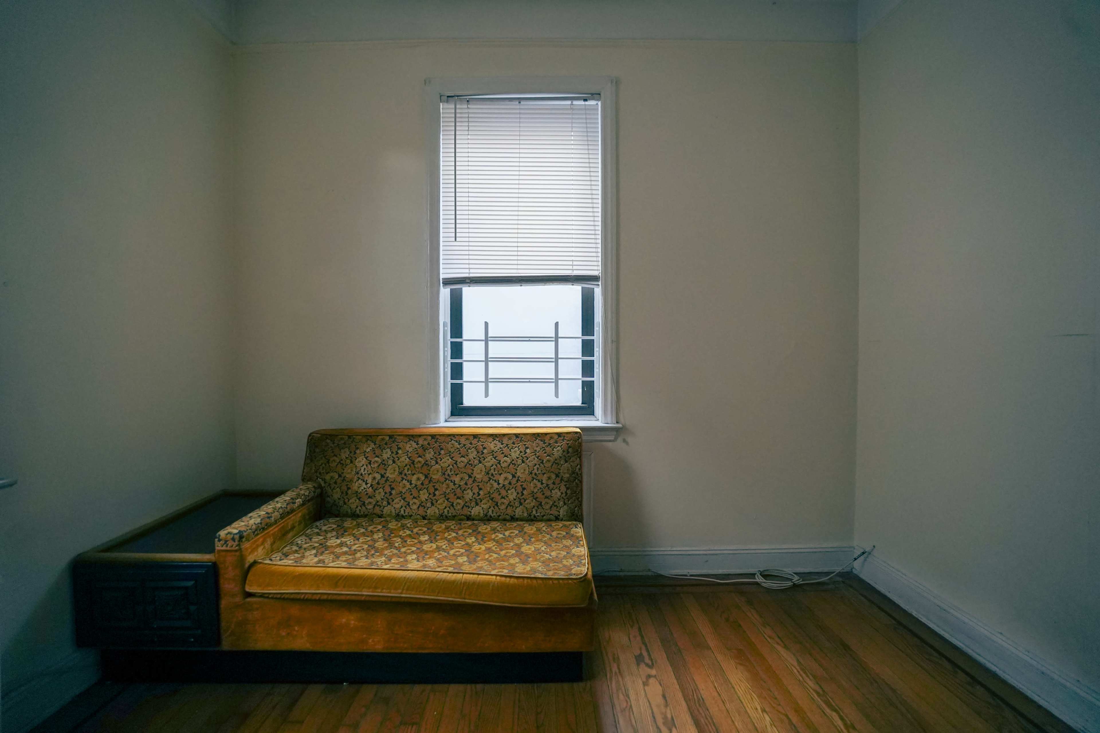 A vintage couch is positioned against a bare wall near a window with closed blinds in a sparsely furnished room.