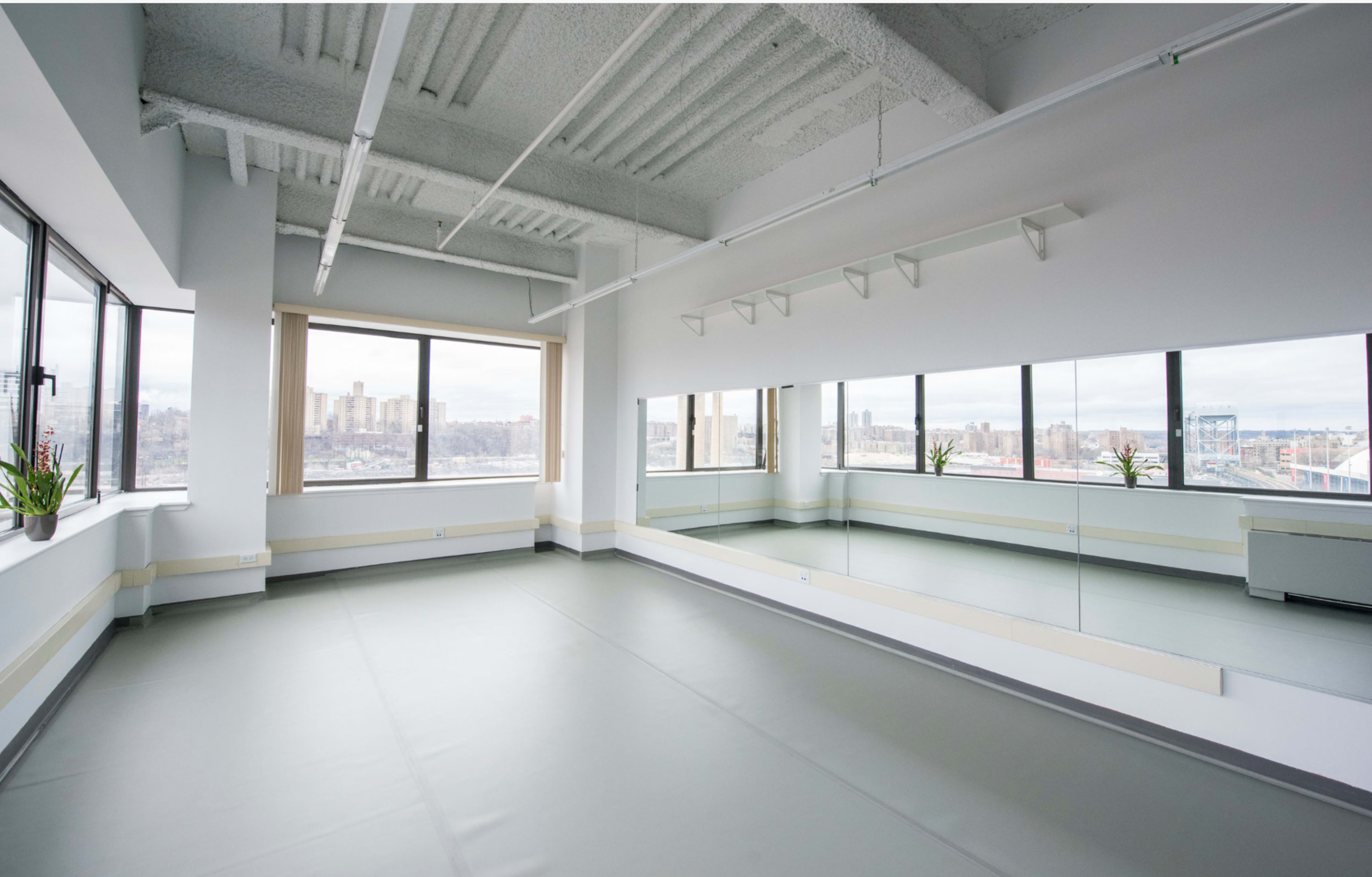 An empty dance studio with large windows, a mirrored wall, and a smooth floor.