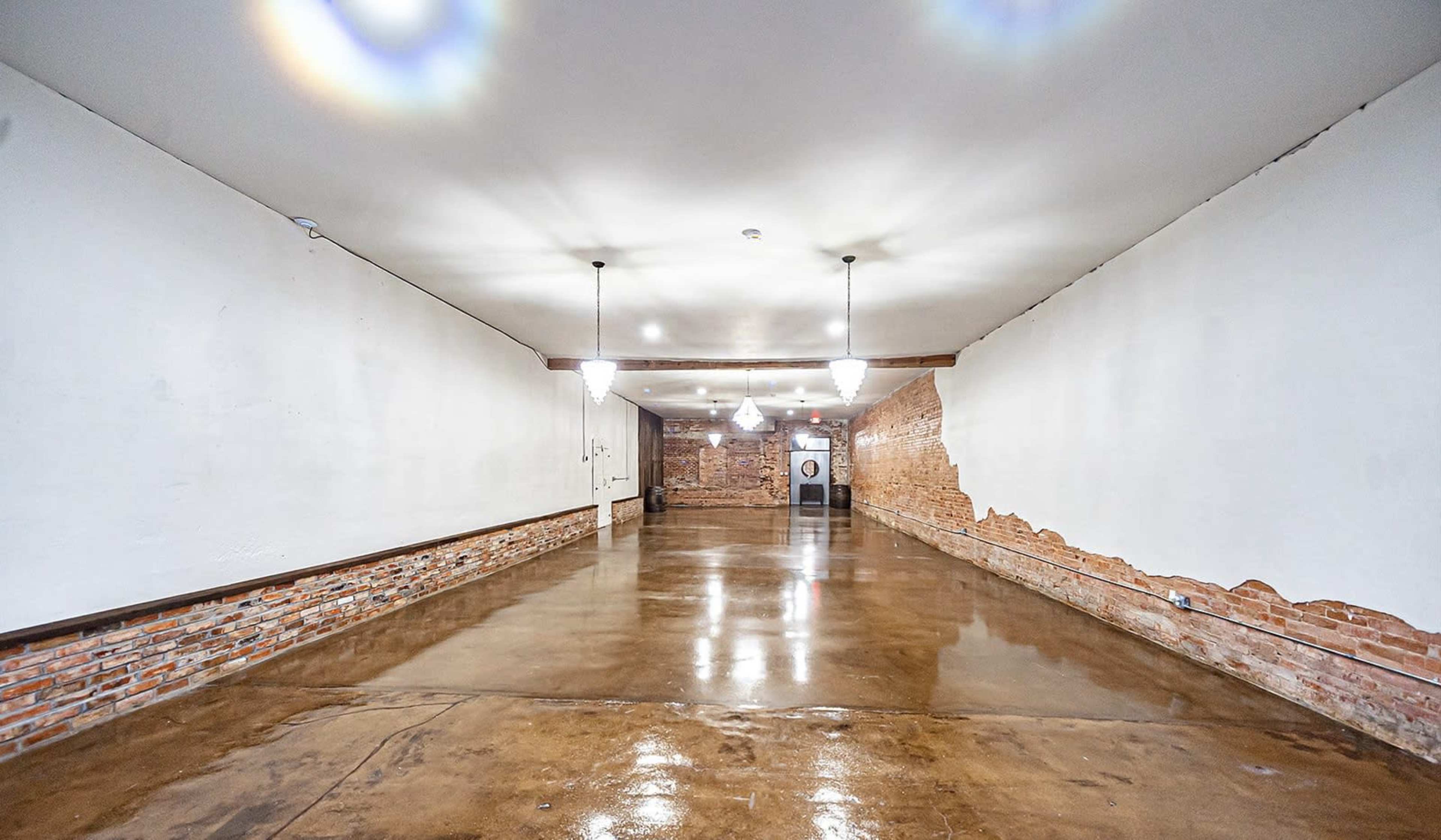 The interior of a spacious room features concrete flooring, exposed brick walls, and several pendant lights hanging from the ceiling.